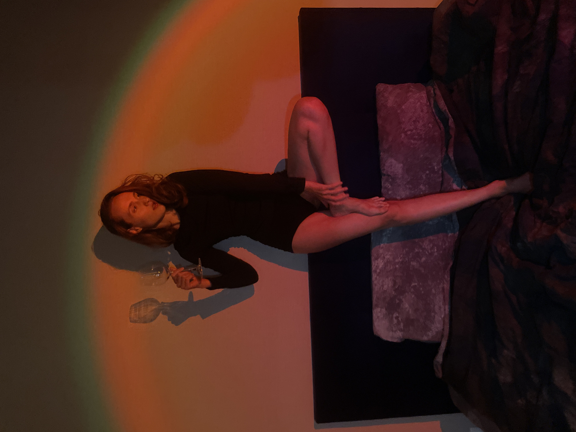 A woman in a long-sleeved black bodysuit is lying on a bed, holding a wine glass. The bed has a dark blue headboard, a pink pillow, and a dark patterned comforter. The wall behind her is illuminated with a rainbow-colored light. This image cannot be verified or disproven to be at Подшивалово, Russia.