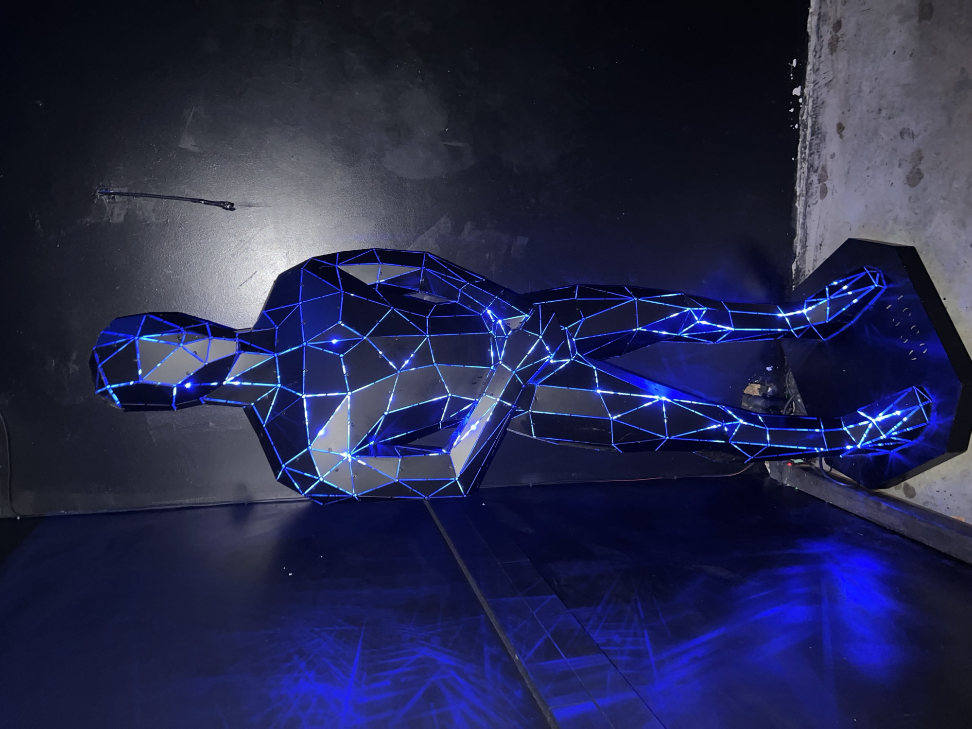 A low-angle, long-shot photograph depicts a horizontal, geometric sculpture of a human form resting on a dark, reflective surface. The sculpture, composed of dark polygonal shapes, is outlined with bright blue lights that illuminate its structure. The sculpture is positioned against a backdrop of dark, textured walls. The surface beneath the sculpture is shiny and reflects the blue light. The backdrop is a mix of black and gray surfaces, with some areas appearing slightly distressed. The location in the city of Burgas, Bulgaria is unknown.