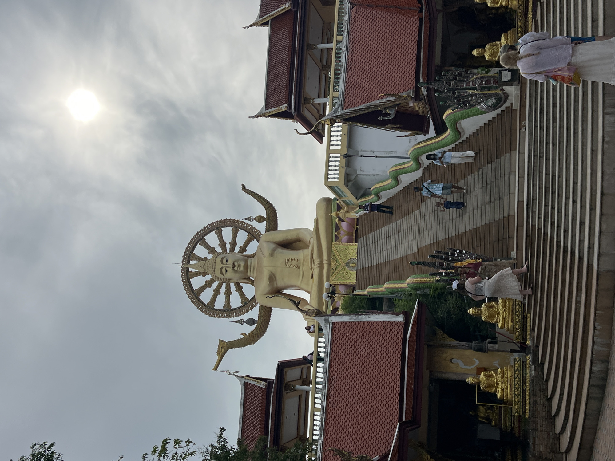 The image shows the Big Buddha of Koh Samui, located at Wat Plai Laem in Baan Plai Laem, Thailand. A massive golden Buddha statue sits atop a tiered structure, with a radiating halo behind its head. In front of the statue, a long staircase with a green serpent-like railing leads down. Several people are visible on the stairs and the platforms, some ascending and some descending. The surrounding buildings have traditional Thai architecture with red tiled roofs and ornate decorations. The sky is overcast with the sun visible as a bright orb.