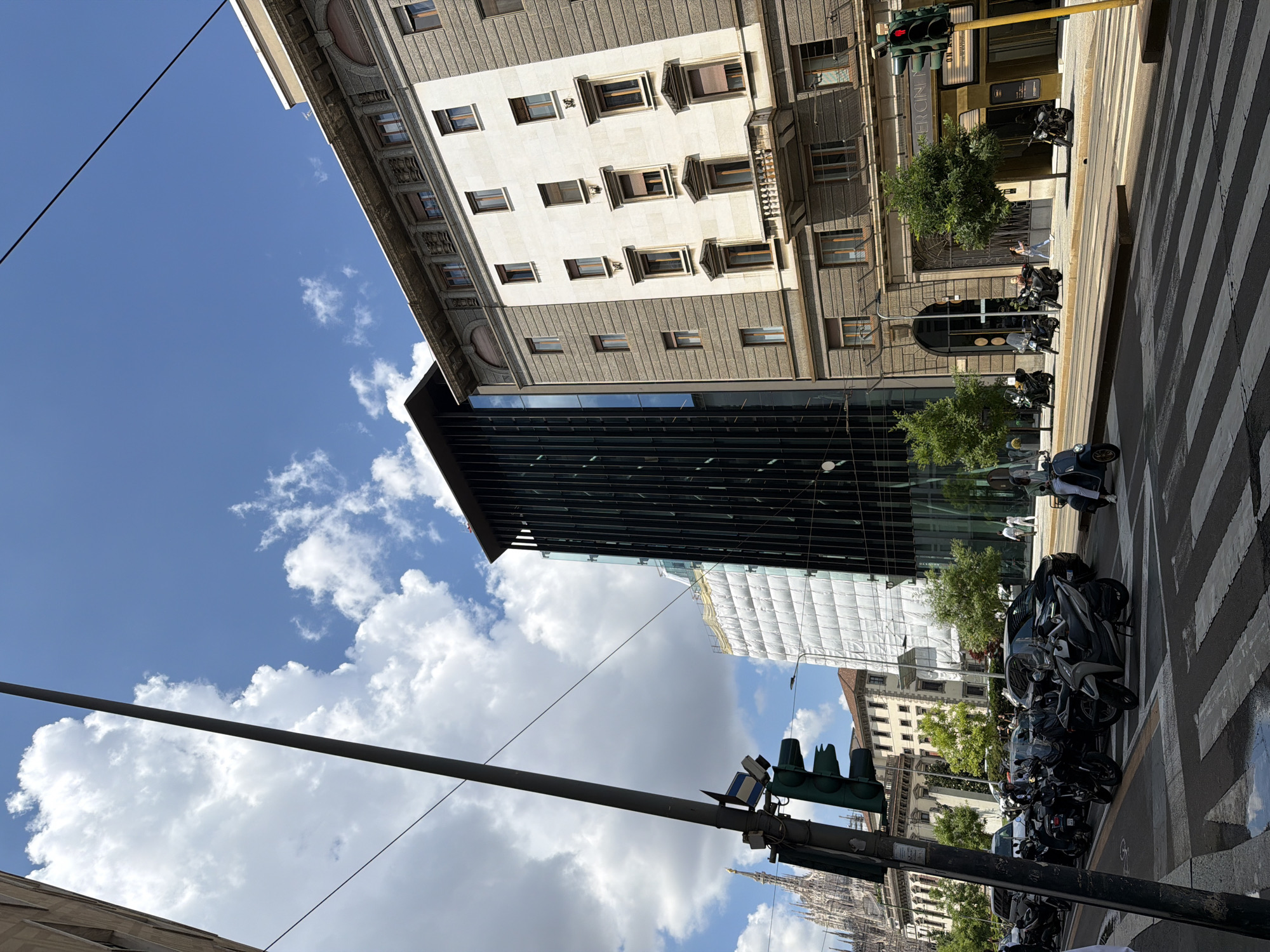 The image presents a vibrant urban street scene in Milan, Italy, captured on a clear, sunny day with a bright blue sky and scattered white clouds. The perspective is looking upwards from street level, showcasing a mix of architectural styles.
To the left, a stately, light-colored historic building with numerous windows, some featuring decorative surrounds and balconies, dominates the frame. Its facade includes sections of darker, possibly stone, elements. Adjacent to this, a modern building with a dark, reflective glass facade rises, reflecting the sky and surroundings. A section of this area appears to be under construction or renovation, with scaffolding draped in white protective sheeting visible.
At street level, a broad pedestrian zebra crossing is prominent. The sidewalks are lined with a multitude of parked motorcycles and scooters, a characteristic feature of Italian cities. Traffic lights on a yellow pole show a red signal, while another set of lights appears green. Small, leafy trees are planted along the pavement, adding a touch of nature to the urban landscape. Several individuals can be seen walking on the sidewalks, appearing relatively small against the imposing buildings. In the distant background, the intricate spire of the Duomo di Milano is faintly visible, confirming the location.
The bright daylight casts clear shadows, suggesting it is mid-morning or early afternoon. A partial sign on one of the older buildings reads "MERCI". The overall scene depicts a bustling cityscape where historical grandeur meets contemporary design and everyday street activity.