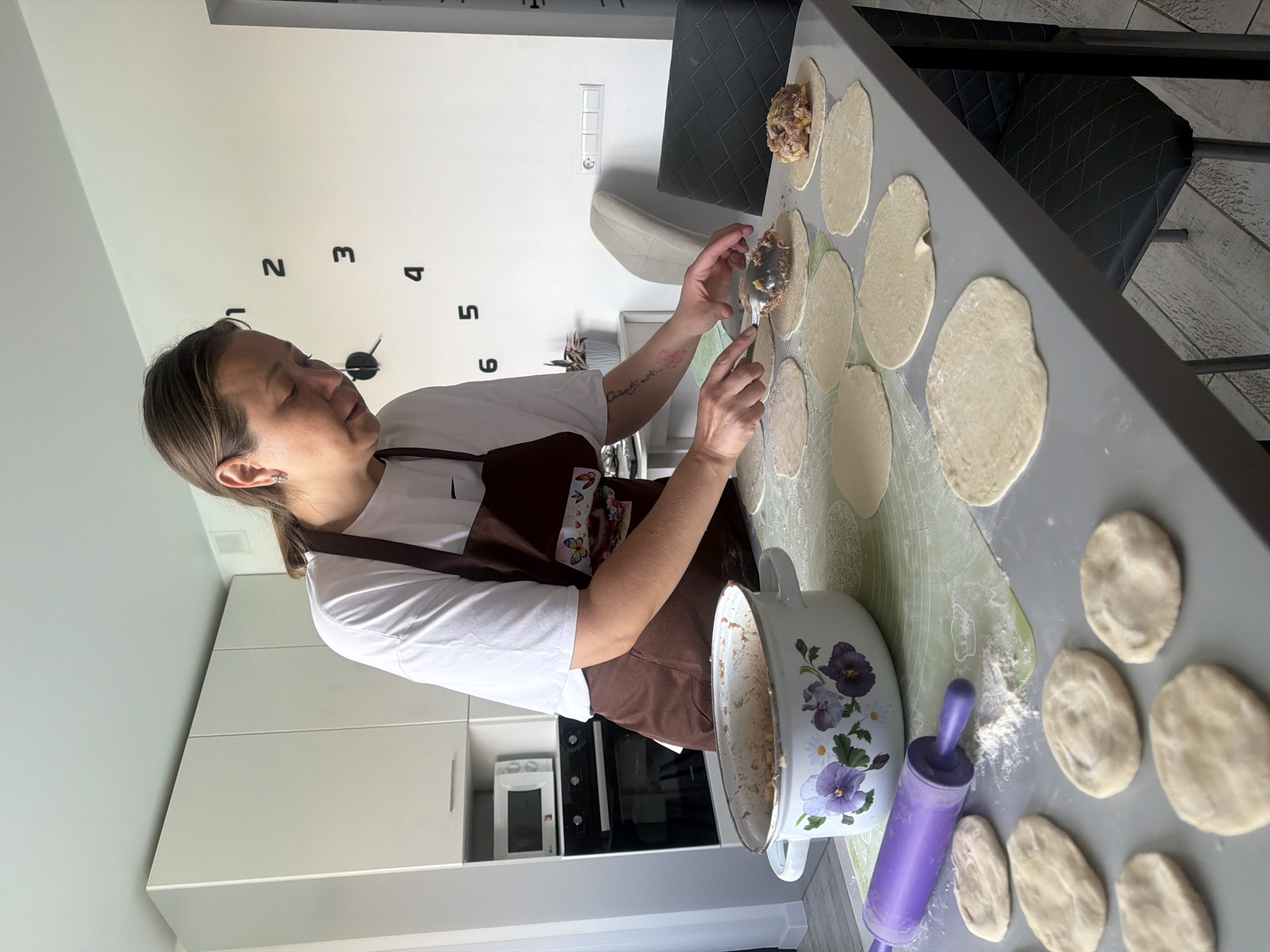 In a brightly lit kitchen, a woman is preparing food. She wears a white t-shirt and a brown apron with a butterfly design. The woman is standing at a gray countertop, where she is using a spoon to place filling onto circles of dough. Several circles of dough are laid out on a light green surface. A large, white pot with floral decorations is next to a purple rolling pin on the counter.
Behind her are white kitchen cabinets with a built-in microwave and oven. A large, decorative clock is on the wall, above the cabinets. A gray chair with a quilted pattern sits next to the countertop.
The location is identified as Chaikovsky, Russia.
