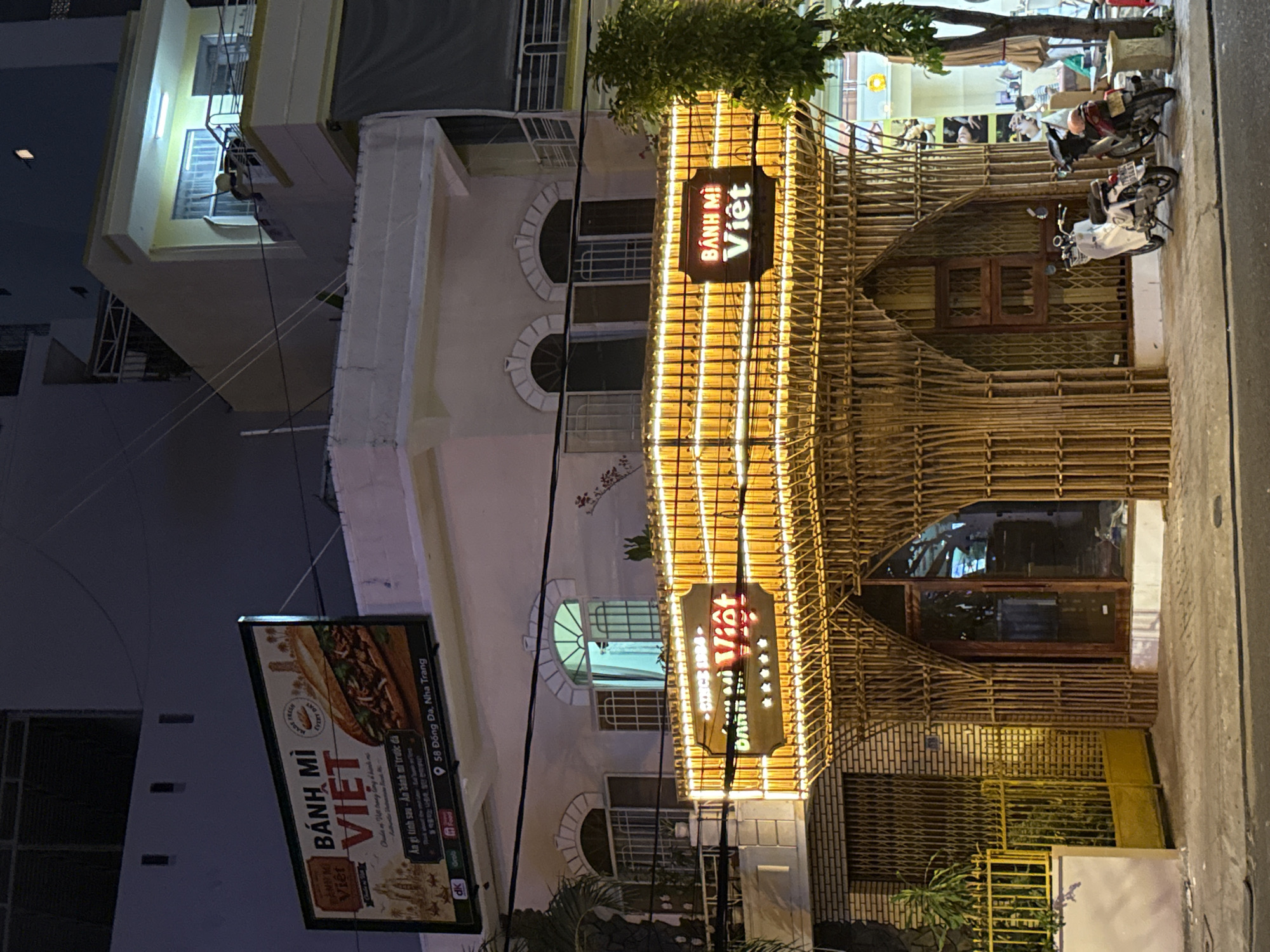 At night in Nha Trang, Vietnam, a storefront with a bamboo facade is illuminated by strings of warm, yellow lights. A prominent sign above the entrance reads "BÁNH MÌ VIỆT," with the words "SINCE 1989" displayed above them. The facade features an arched entryway made of bundled bamboo poles. In front of the store, two motorcycles are parked on the sidewalk. To the left of the entrance, a yellow metal gate is visible. Above the storefront, a large rectangular billboard advertises "BÁNH MÌ VIỆT" with an image of a sandwich. The billboard also displays an address: "58 Đông Da, Nha Trang." The surrounding environment suggests a city street scene, with buildings and power lines visible in the background.