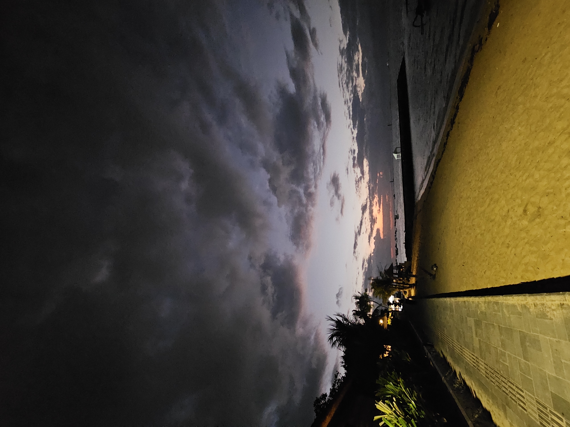 The image depicts a twilight scene on a beach in Denpasar, Indonesia. A paved walkway runs along the left side of the frame, bordering a sandy beach. The sky is filled with dark, dramatic clouds, with hints of pink and orange near the horizon, suggesting either sunrise or sunset. The ocean is visible to the right, with a dark structure extending into the water. Palm trees and vegetation are present on the left side, with some artificial lighting illuminating the area. There are no people or specific actions visible in the image.