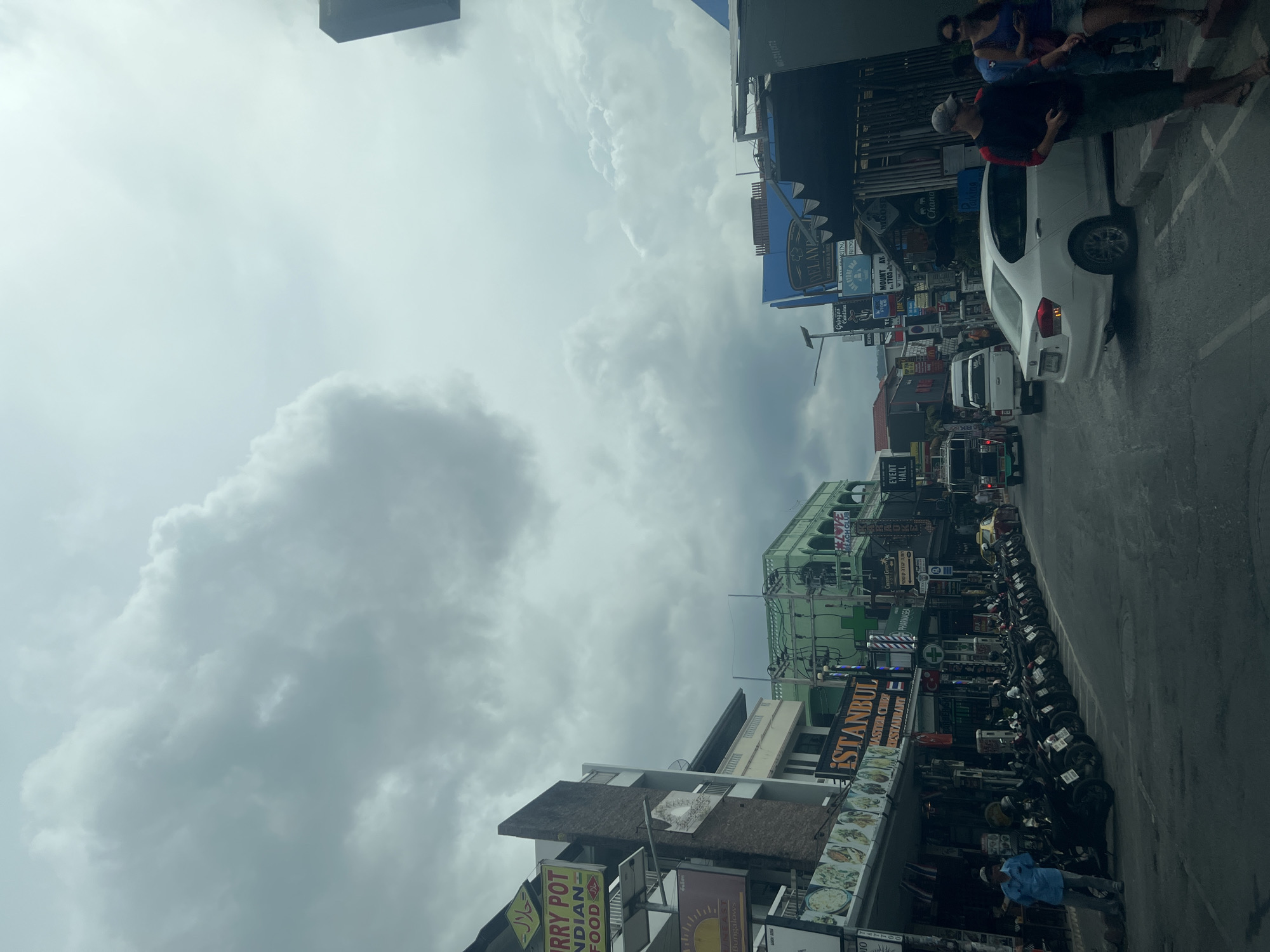 The image shows a street scene in the Ko Samui District of Thailand under an overcast sky. The street is lined with buildings featuring various signs, including "CURRY POT INDIAN FOOD," "ISTANBUL MASTER CHEF RESTAURANT," "PHARMASEA," and "EVENT HALL." A row of parked motorcycles is visible along the side of the road. Several people are present, including a man in a blue shirt standing near the motorcycles, and two individuals, a man and a woman, walking on the sidewalk. A white sedan is driving on the street, and other vehicles are also visible in the background. The environment appears to be a commercial or tourist area with a mix of shops and restaurants.