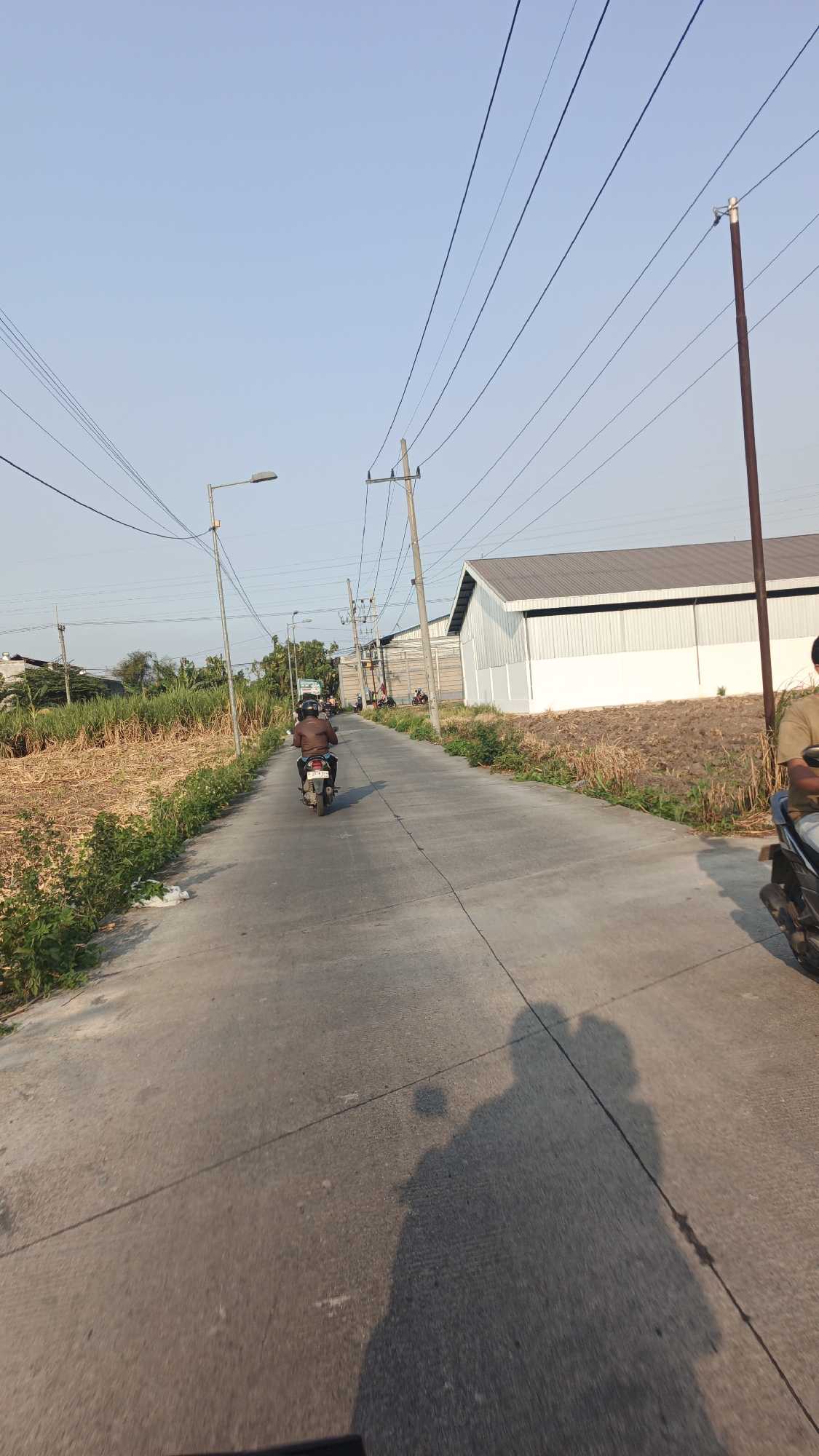 The image depicts a paved road with a single rider on a motorcycle in the foreground, traveling away from the viewer. The rider is wearing a brown jacket and a helmet. To the left of the road, there is dry vegetation and some green plants along the roadside. In the background, there is a long, white building with a metal roof, likely a warehouse or industrial facility, characteristic of the industrial areas found in Sumolawang, Indonesia. Utility poles with power lines are visible stretching across the sky, and a street lamp is present on the left side of the road. The sky is clear and blue, suggesting daytime. Shadows of the motorcycle and its occupants are cast on the road, indicating the direction of sunlight. There are other indistinct figures on motorcycles further down the road.