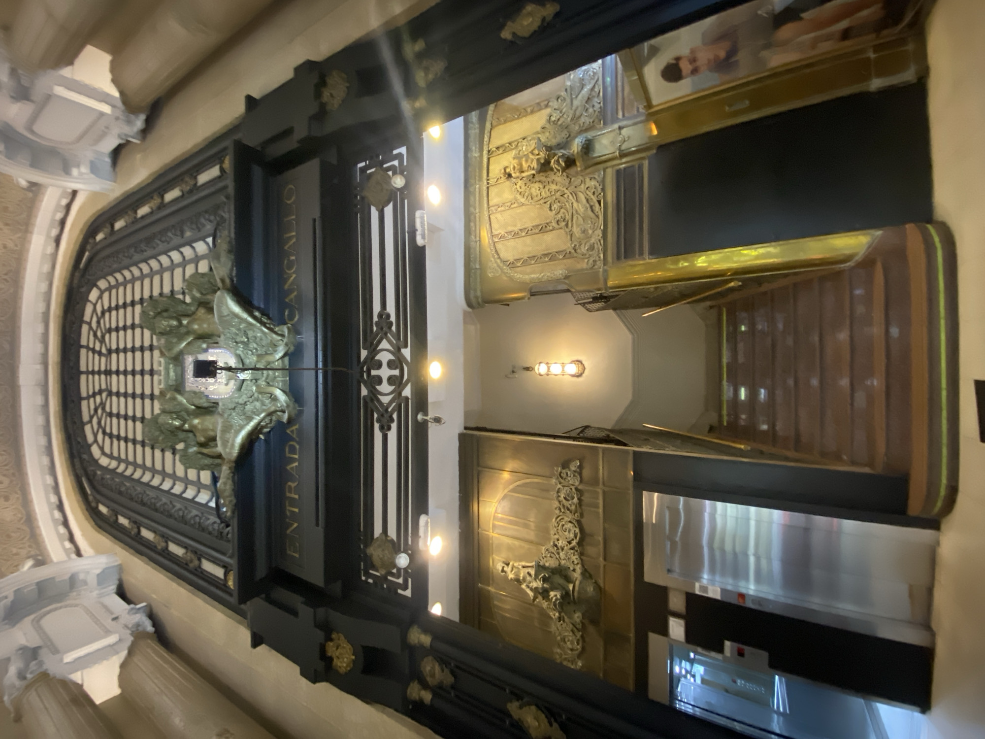 This is an interior shot of the entrance to Galeria Güemes, located at Florida Street 165 in Buenos Aires, Argentina. The view appears to be taken from the mezzanine level looking downwards. The architectural details include decorative ironwork and gilded ornamentation. The main features are the entrance doors with "Entrada" and "Cangallo" signs on their pillars. Further inside, the image reveals a staircase and a glimpse of the architectural details beyond the lobby. The environment is artificially lit. No people are visible.