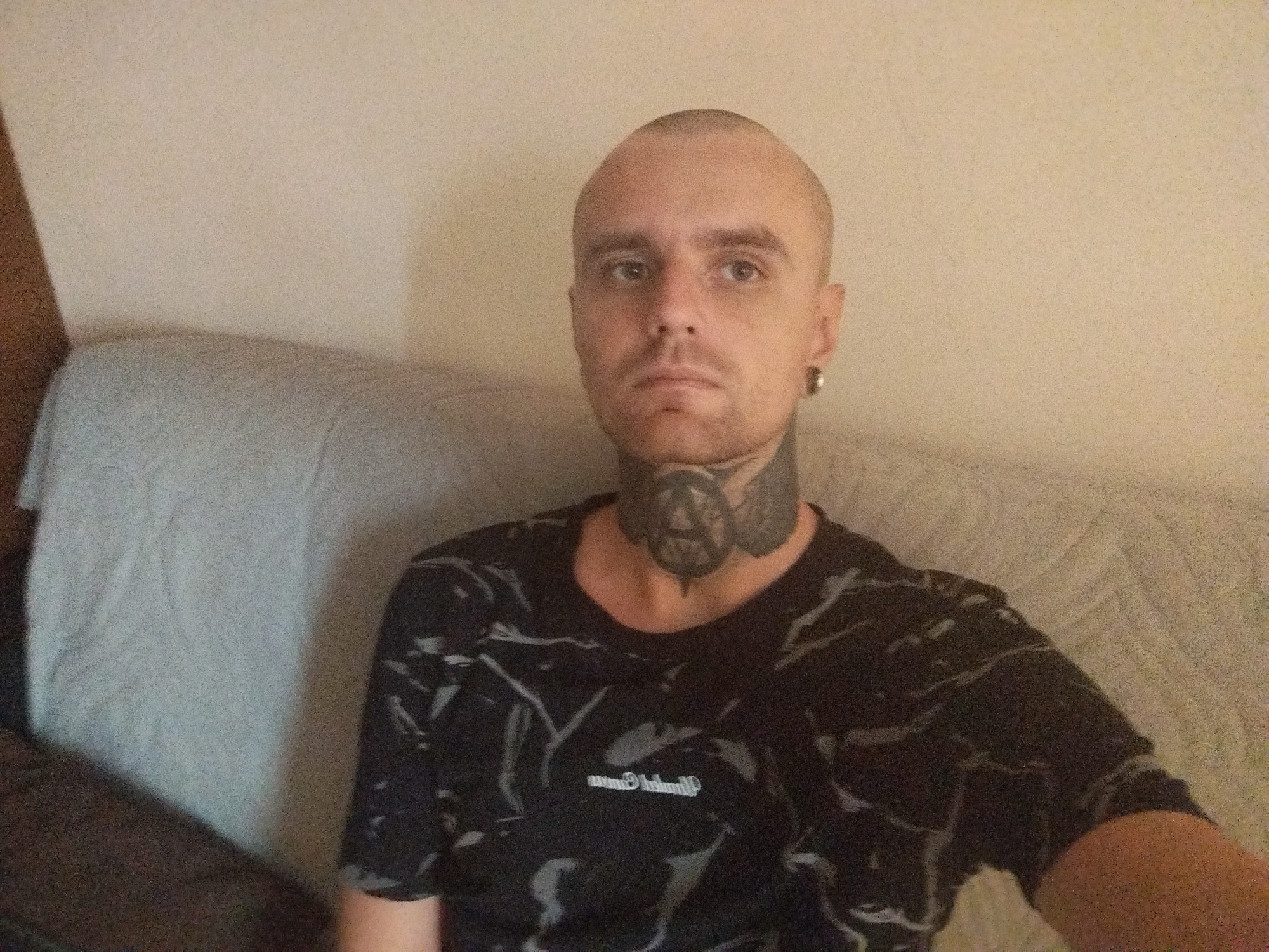 A man with a shaved head and tattoos on his neck is sitting on a couch in what appears to be a living room. He is wearing a black t-shirt with a white abstract pattern. The couch is covered with a light gray blanket. The wall behind him is a neutral beige color. The man is looking directly at the camera with a neutral expression. He has a silver hoop earring in his left earlobe. The lighting in the room is dim. There is no specific indication of location in Batumi, Georgia, within the image itself.