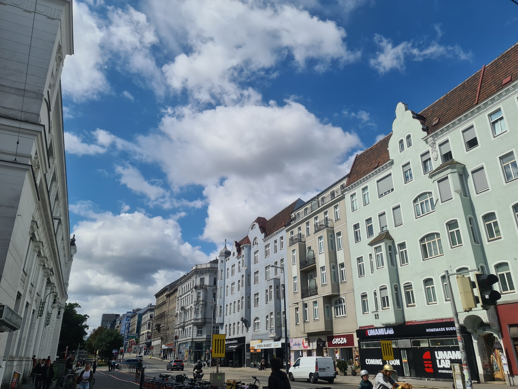 The image depicts a street scene in Vienna, Austria, with a focus on the architecture of the buildings lining the street and a bright, cloudy sky. The street is lined with multi-story buildings featuring a variety of architectural styles, from classical facades to more modern designs. The buildings are painted in different colors, including white, light blue, and a pale green. Many of the buildings have tiled roofs, and some feature balconies and numerous windows.
On the street level, there are various shops and businesses with signs. Some of the visible signs include "UMAMILAB," "HOMEMADE BURG," "PARSA MARKET," and "GYOZA." A white van is parked on the street, and several bicycles are visible, suggesting pedestrian and bicycle-friendly infrastructure. There are a few people walking on the sidewalk and riding bicycles, all appearing to be going about their daily activities. Traffic lights are present, indicating a regulated intersection. The sky is a vibrant blue with scattered white clouds, suggesting a pleasant day. The overall impression is that of a typical urban street in a European city, with a mix of historical and contemporary elements.