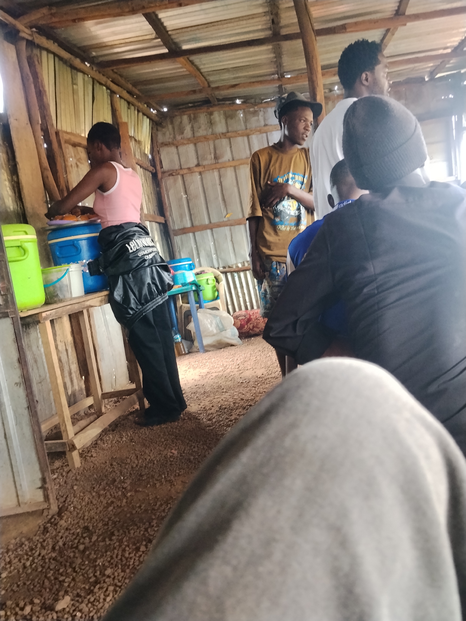 In the city of Unknown, Unknown, within a rustic, open-air establishment with a corrugated metal roof and wooden supports, a group of individuals are gathered. In the foreground, a person's lower body is visible, clad in light grey fabric. Further into the scene, a woman wearing a pink top and black pants stands near a table laden with containers, appearing to be serving food. She has a black jacket with white lettering draped over her arm. Behind her, three men are present. One man, wearing a brown t-shirt and a hat, stands to the left, looking towards the center. Another man in a white t-shirt is positioned behind him, and a third man wearing a black beanie is closest to the foreground, facing away from the camera. The environment is characterized by dirt and gravel flooring, with various containers and objects scattered around the serving area.