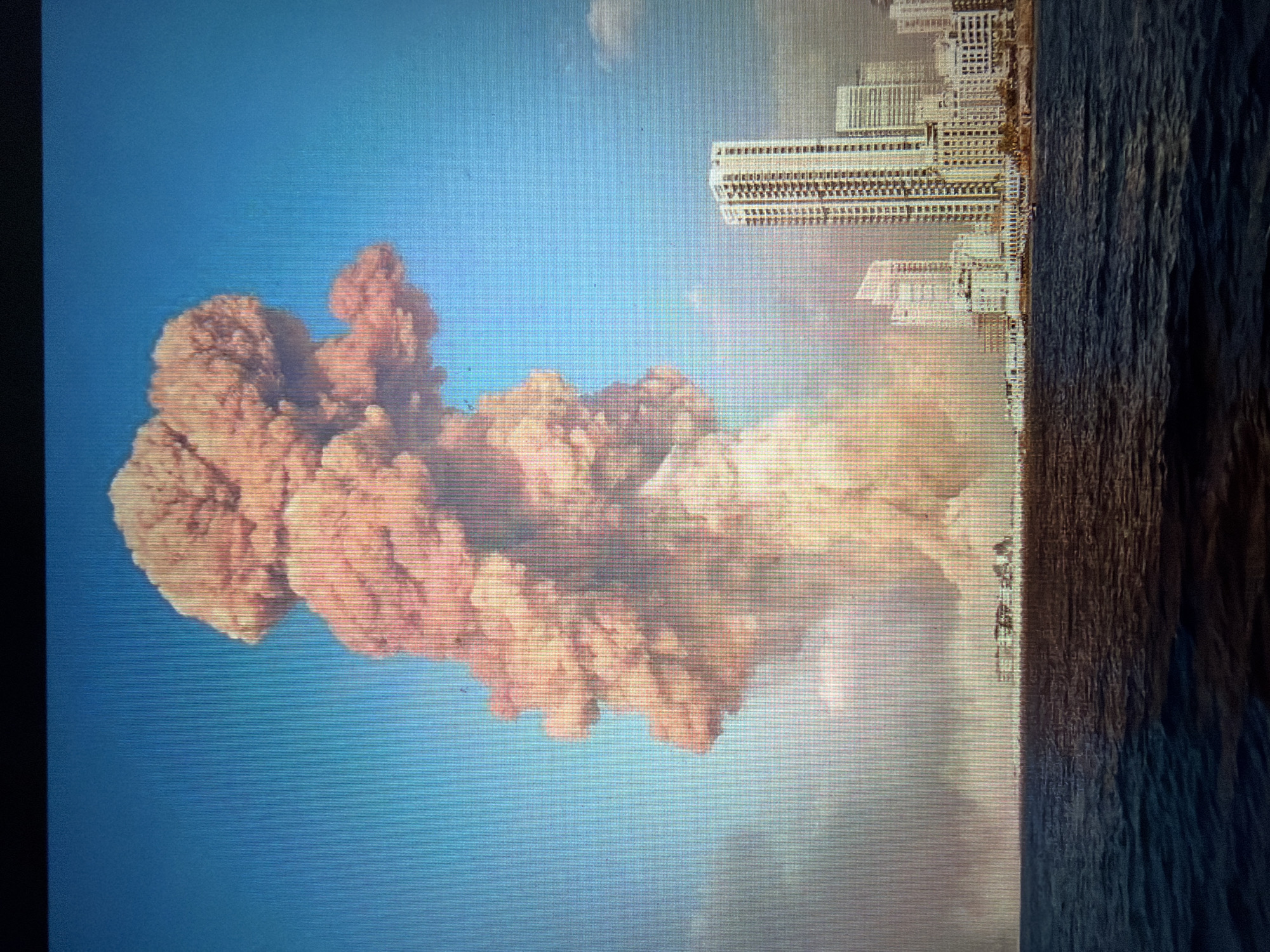 The image depicts a large, pinkish-orange cloud of smoke or debris rising into a clear blue sky. The cloud is voluminous and appears to be expanding, with a textured, turbulent surface. To the right of the cloud, a cityscape is visible, featuring several tall, modern buildings with many windows. The buildings are a light color, possibly white or beige. Adjacent to the cityscape is a body of dark, choppy water, likely the sea, with visible waves. The scene appears to be set during the daytime, given the bright sky. The overall impression is one of a dramatic event, possibly an explosion or a large-scale incident, occurring near a coastal city. There is no visible text in the image. The composition suggests a powerful, impactful moment.