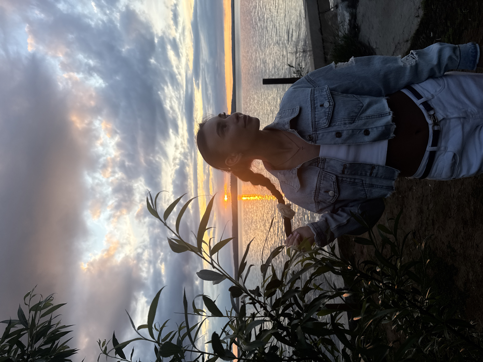 A vertical medium shot shows a young woman with her head tilted upward as she looks toward the sunset over the water in Chaikovsky, Russia. She stands near a tree that has leaves that are the same shade as her hair. She has her hair in a braid and is wearing a denim jacket, a white top, and white pants. The water is in the background, and the sky is filled with clouds.