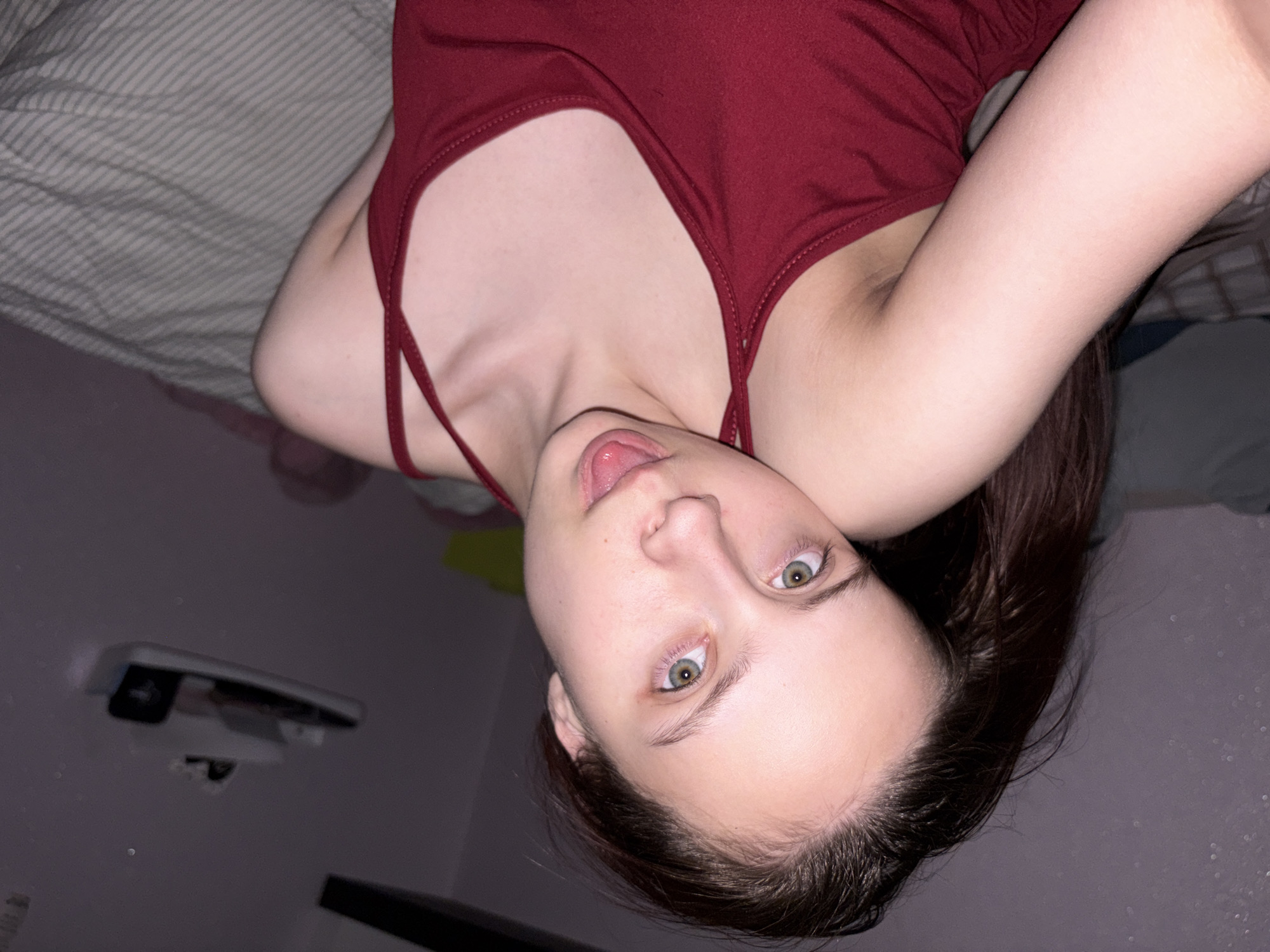 A young woman lies on a bed in a room. She is wearing a red tank top. The bed has a striped grey cover. A partially visible white object is mounted on the wall in the background. The location is stated as Прикамский, Russia.