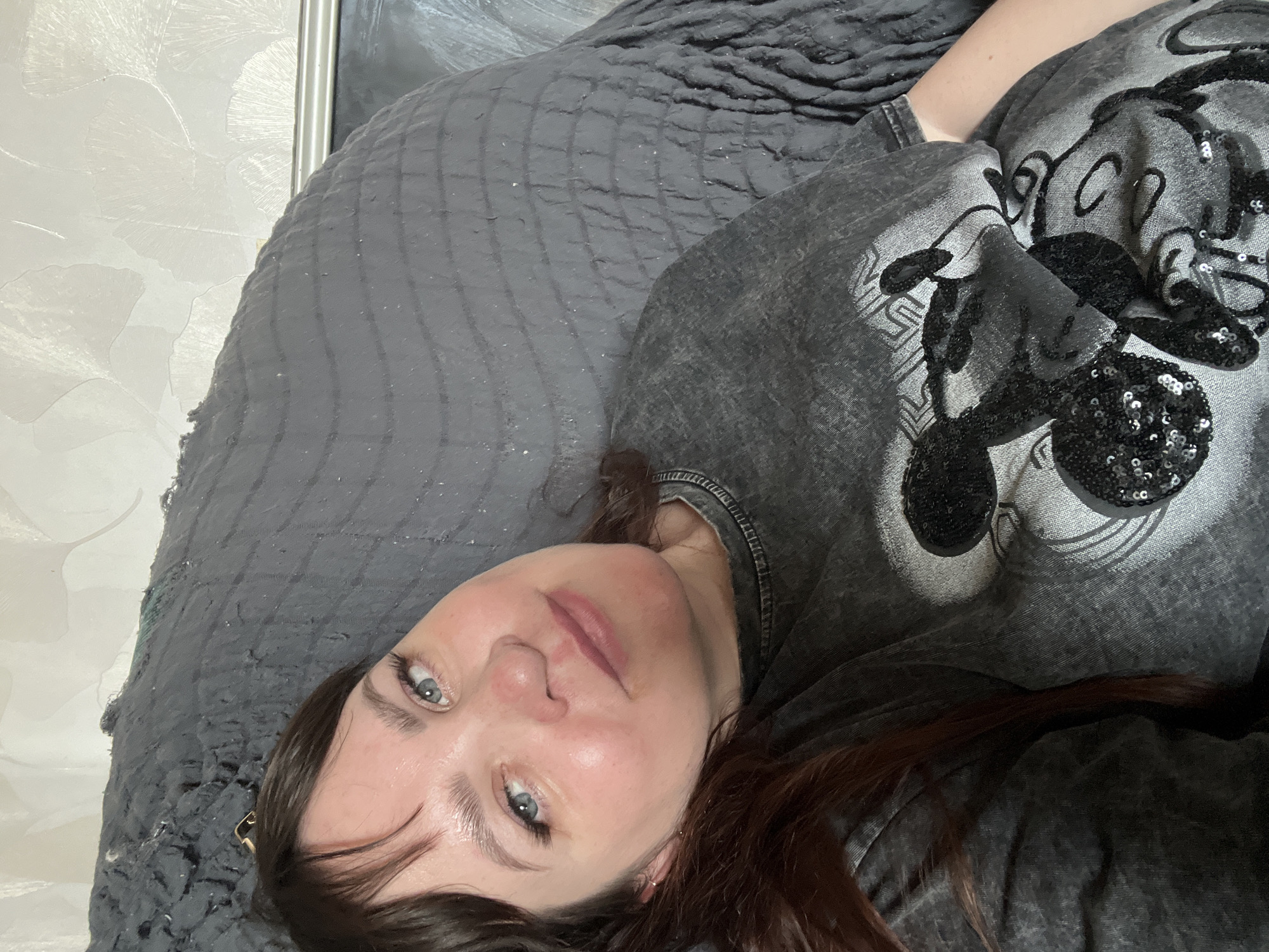 A woman with brown hair is lying on a dark grey quilted bedspread. She is wearing a grey t-shirt with a Mickey Mouse design embellished with black sequins. The woman's face is centered in the frame; she has light skin, blue eyes, and pink lips. The background includes a patterned wallpaper in light shades. This image was not taken in the city of Chaikovsky, Russia.