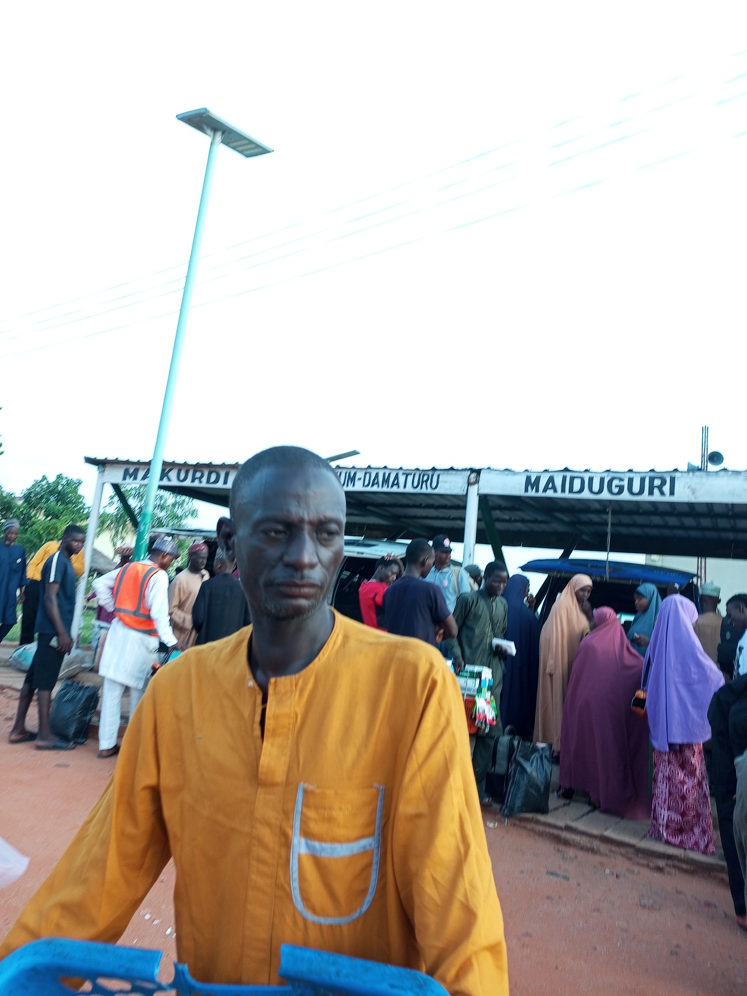 The image depicts an outdoor scene at a location in Gombe, Nigeria, likely a transportation hub or market, given the presence of a sign displaying "MAIDUGURI" and "MAKUDI". In the foreground, a man with dark skin and a close-shaved head is prominently featured, wearing a bright orange tunic with a light blue trim on the chest pocket. He is looking towards the camera with a neutral expression. Behind him, a diverse group of people are gathered. Some are dressed in traditional attire, including women wearing headscarves and long dresses in various colors like purple, pink, and beige. Men are seen in a mix of casual wear and what appears to be work attire, such as a person wearing an orange safety vest over a white garment. Several black plastic bags and other items are scattered on the ground, suggesting a busy environment with people possibly traveling or engaged in commerce. A tall, light blue pole with a solar-powered lamp at the top is visible on the left side of the image, indicating an urban or semi-urban setting. The ground is a reddish-brown earth, and the sky is overcast and white.