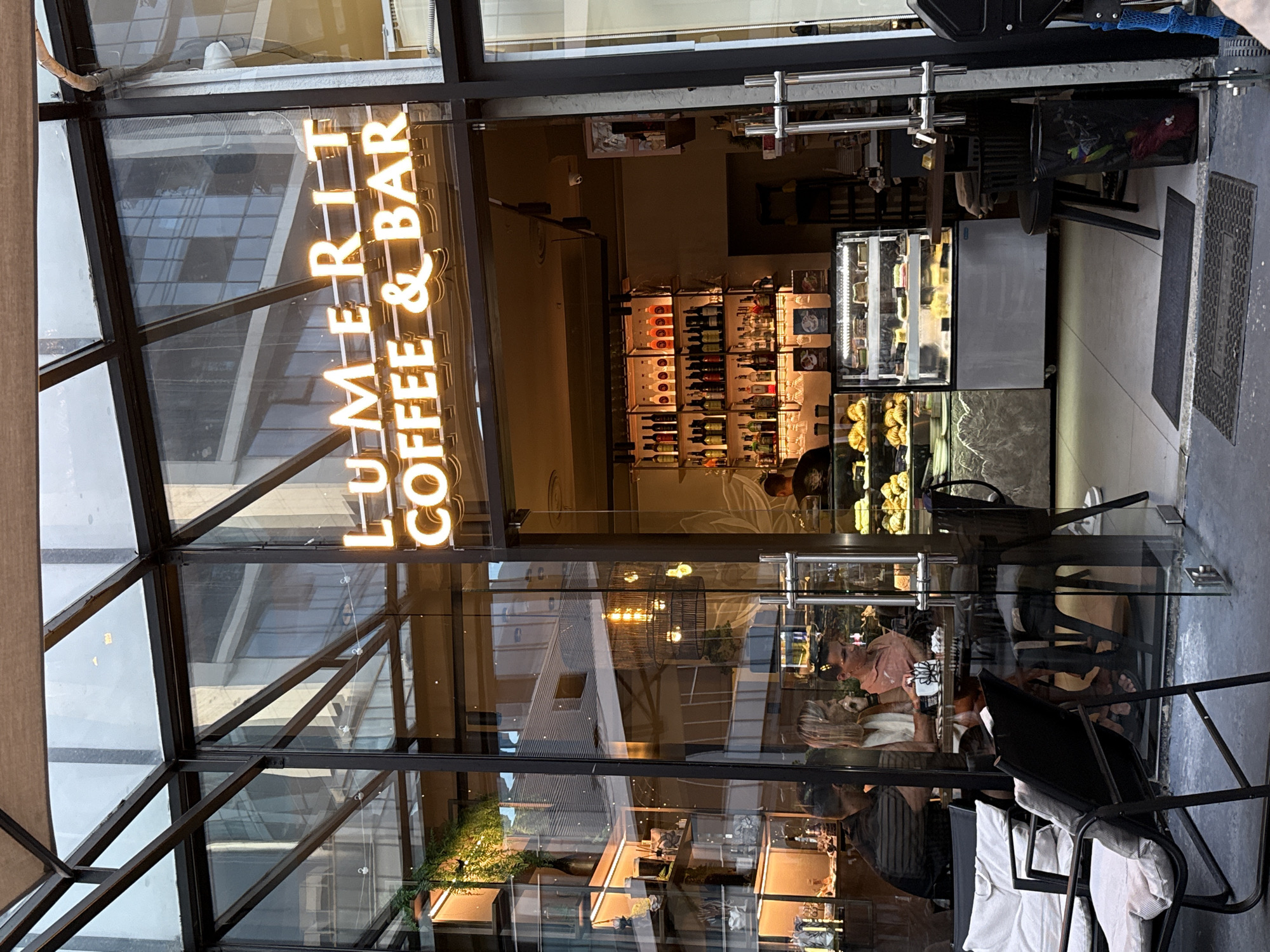 The image shows the exterior of a coffee shop with a neon sign that reads "LUMERIT COFFEE & BAR". The shop has a glass facade, and inside, shelves are stocked with bottles of alcohol. A display case showcases pastries and cakes. Several people are seated at tables inside the coffee shop, appearing to be customers. Outside the shop, there are chairs and tables, suggesting an outdoor seating area. The environment appears to be urban, possibly a street or an outdoor plaza. The lighting is warm, indicating either interior lighting or evening light.