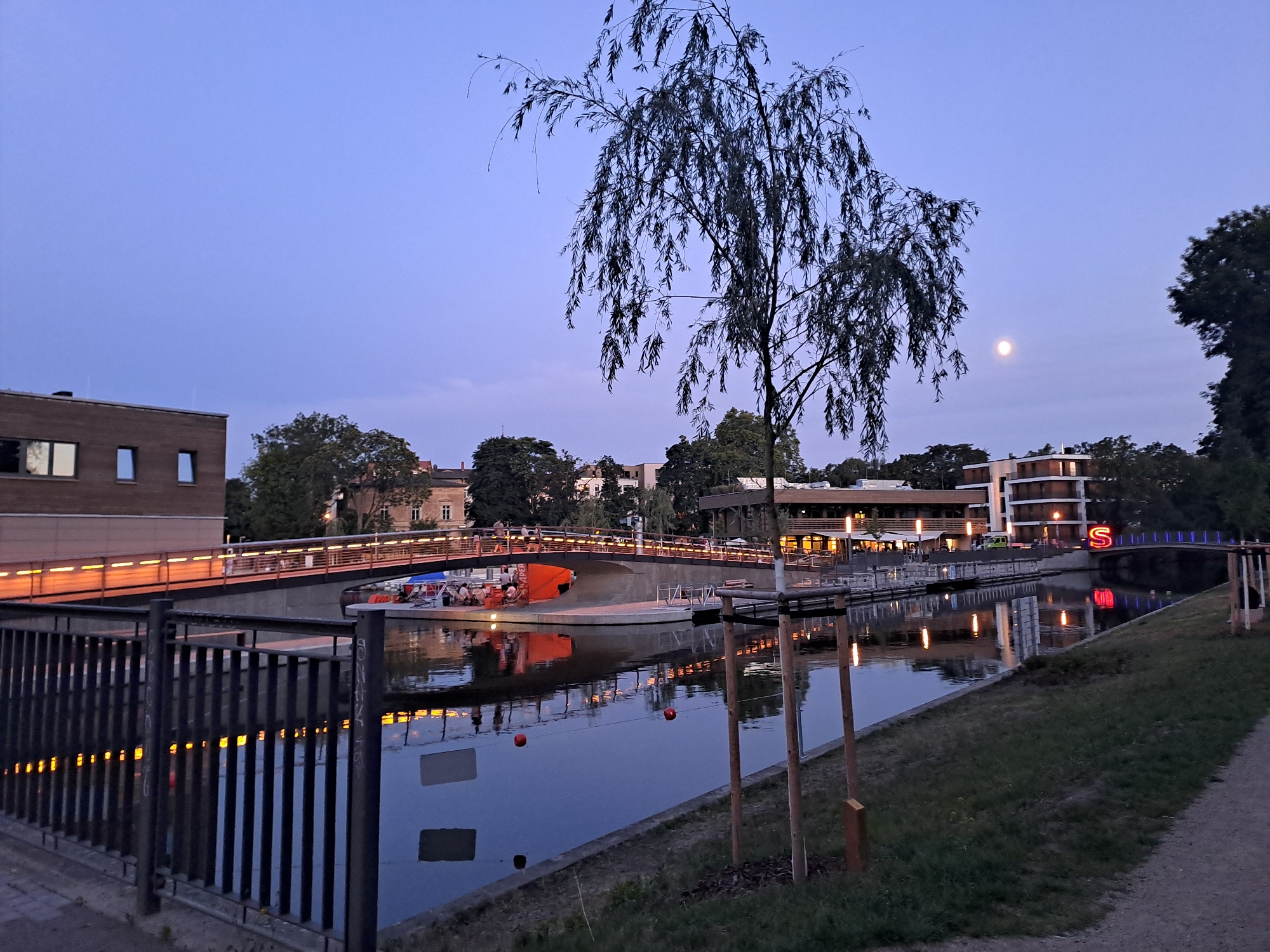 This is a night-time photograph of the River Elster in Leipzig, Germany, specifically near the new bridge on the Karl-Tauchnitz-Ufer. The image captures the twilight atmosphere with a clear, pale blue sky and the moon visible in the upper right quadrant.
In the foreground, a black metal fence with vertical bars runs along the left side of the frame. The fence casts reflections of warm yellow lights onto the canal water. The water itself is dark and still, reflecting the lights from the bridge and the buildings. Two red buoys are visible floating in the canal. On the right side of the canal, a grassy embankment leads to a dirt path. Several young trees, supported by wooden stakes, are planted on the embankment.
A modern pedestrian bridge spans the canal. The bridge is illuminated from underneath with a continuous line of warm yellow lights, casting a glow on its underside and reflecting into the water. Several people are seen walking and standing on the bridge. Below the bridge, a large orange boat or barge is docked. There are people seated on the deck of this vessel.
On the far bank of the canal, a series of modern buildings are visible. These buildings feature large windows and illuminated interiors, with some displaying a warm orange glow. One building on the right has a prominent red "S" neon sign. Trees line the bank between the buildings. A weeping willow tree with long, slender branches dominates the central and upper left portions of the image. The branches hang down, creating a silhouette against the twilight sky.