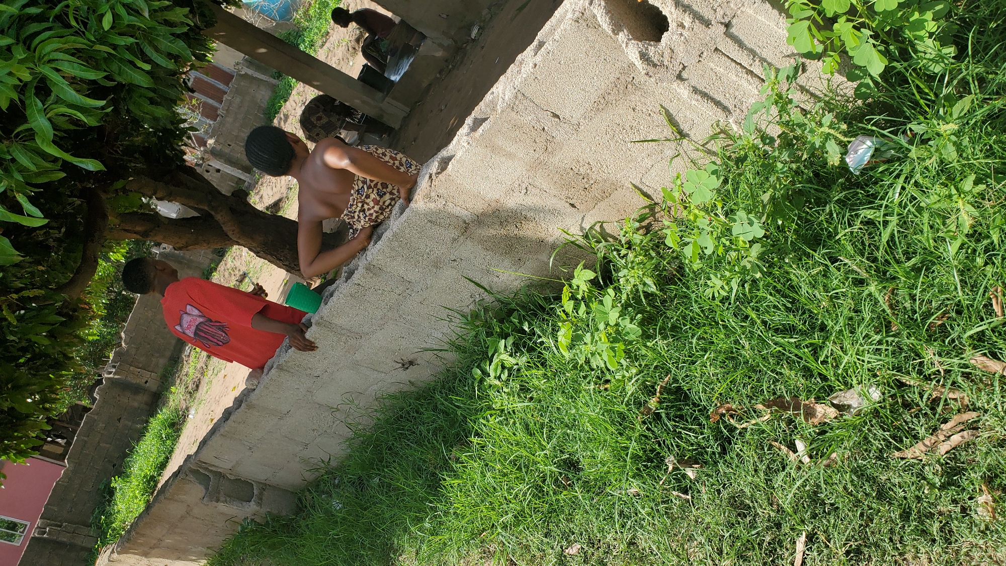 In Jalingo, Nigeria, two individuals are observed near a concrete wall with a tree providing shade. One person, wearing a red t-shirt, leans on the wall, with a green plastic cup visible beside them. The other person is seated on the wall, facing away from the camera, wearing a patterned skirt. In the background, a building with a red facade is partially visible, along with other structures and trees. The ground is covered in grass and some dirt.