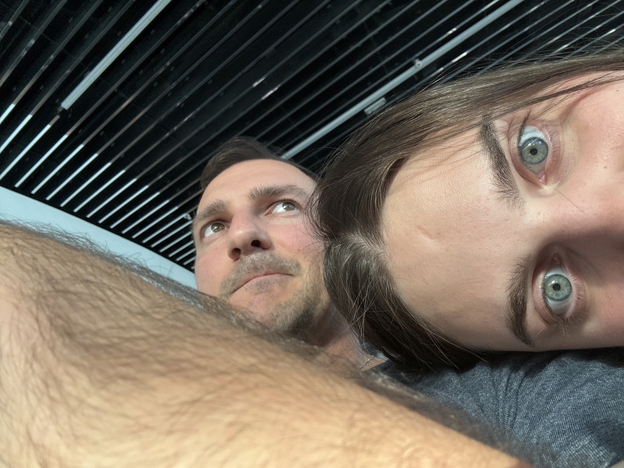 A close-up selfie shows two people and part of an arm. The man is positioned on the left, looking upward with a somewhat thoughtful expression. The woman is positioned on the right, her face tilted at an angle. Part of a hairy arm is visible in the foreground, partially obscuring the subjects. Overhead is a ceiling structure composed of a series of parallel black beams with light strips in between. The photo was likely taken indoors. The location in the city of Schwechat, Austria is not evident in the image.