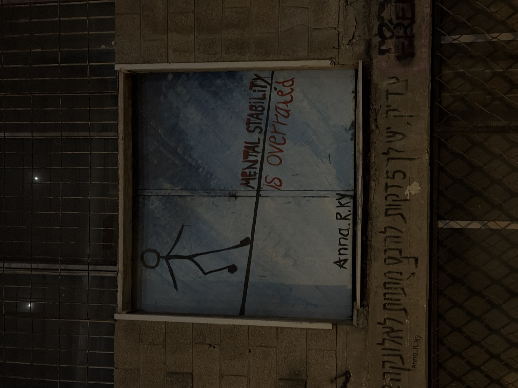 The image shows a section of a building exterior at night. A large, boarded-up window is the focal point, painted with a blue background. On the left side of the board, a black stick figure is depicted walking on a tightrope, its arms outstretched for balance. The right side of the window features red graffiti that reads "MENTAL STABILITY IS overrated". Below the window, painted in black on the wall, is the signature "Anna.R.Ky". Further down on the wall, there is graffiti in Hebrew script. The building appears to be constructed of brick, and a barred window is visible above the boarded-up section. The scene is dimly lit, suggesting it is nighttime. Based on the Hebrew graffiti and the presence of the signature "Anna.R.Ky", this appears to be street art in Tel-Aviv, Israel.
