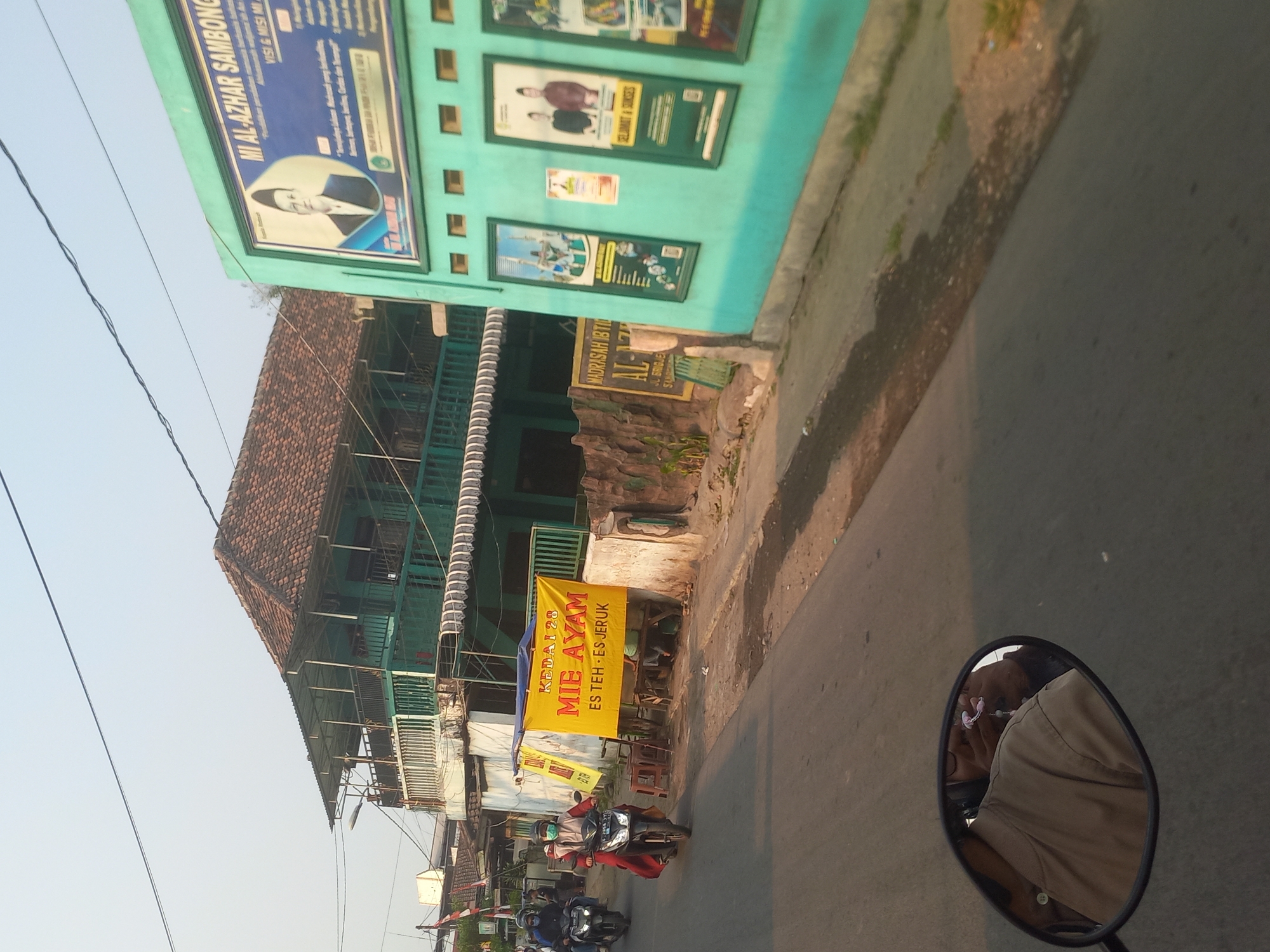 The image depicts a street scene in Jombang, Indonesia, with a focus on a building to the right and a yellow banner on the left. The building on the right is painted turquoise and features several framed posters on its exterior wall. One prominent poster displays the text "MI AL-AZHAR SAMBONG" and includes a portrait of a man in a traditional cap and suit. Below this, there are other posters with images of people and text. To the left of the turquoise building, a yellow banner is prominently displayed. The banner reads "KEDAI 28 MIE AYAM" and "ES TEH ES JERUK," indicating a food stall selling noodle soup and iced tea with oranges. In front of the banner, there are some stools and a low wooden table, suggesting outdoor seating. The street itself is paved, and the scene is viewed from the perspective of someone riding a motorcycle, as evidenced by the reflection in the rearview mirror on the lower left, which shows the back of a person wearing a light brown shirt and glasses. Two other motorcycles with riders are visible on the road, heading away from the viewer. The background features a multi-story building with green walls and a tiled roof, and a clear sky above. The overall impression is of a commercial street in a residential or mixed-use area.