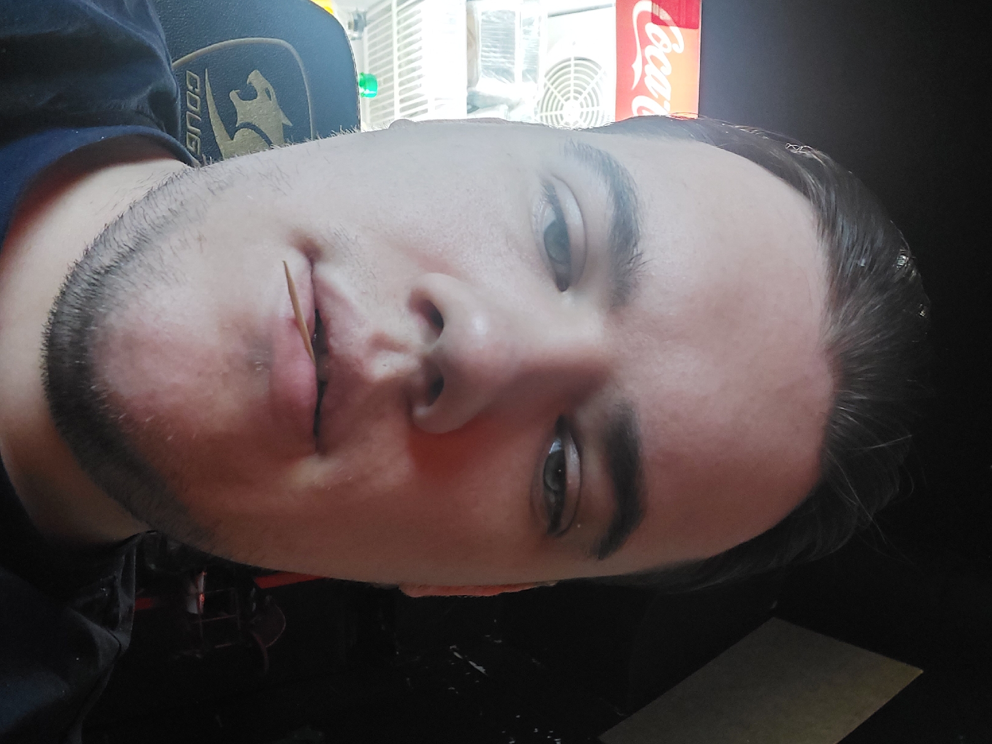 A close-up portrait of a light-skinned male with short dark hair and a beard, holding a toothpick in his mouth. He is wearing a dark-colored shirt with a logo partially visible on the shoulder. The background includes a Coca-Cola advertisement and what appears to be a refrigerator unit. The location is not evident based on the visual information in the image.