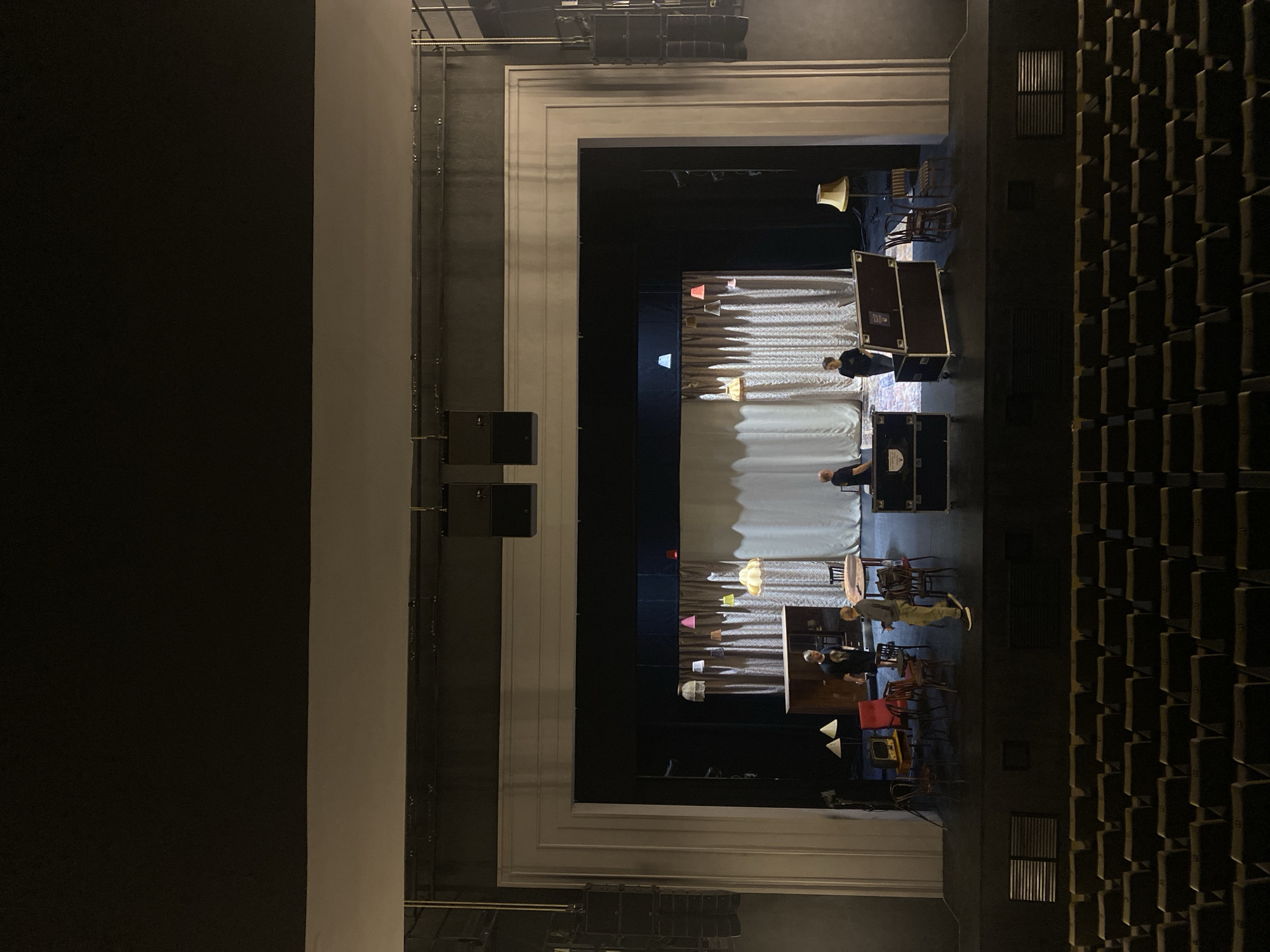 The image depicts an empty theater auditorium, likely located in Elino, Russia, based on the prompt. The stage is visible through the proscenium arch. Two men appear to be setting up props, including furniture, curtains, and lighting fixtures. Equipment cases are present. The seating area is dark and unoccupied. Speakers are mounted on the sides of the proscenium.