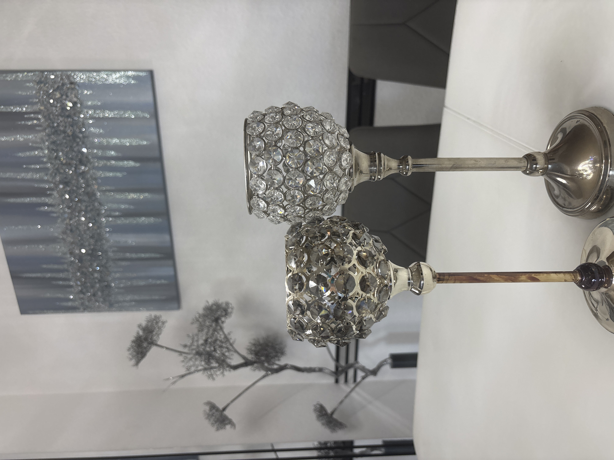 Two crystal-encrusted candle holders stand on a white surface. The candle holder on the left is slightly shorter and has a stem with a mottled brown and gold appearance. The candle holder on the right is taller and has a sleek silver stem. Behind the candle holders, a framed artwork with a sparkly, abstract design hangs on a light-colored wall. To the left of the candle holders, a metallic tree-like sculpture with delicate branches is visible. In the background, the backs of two dark gray chairs are partially visible. The scene is indoors, suggesting a home or a decorative display area.