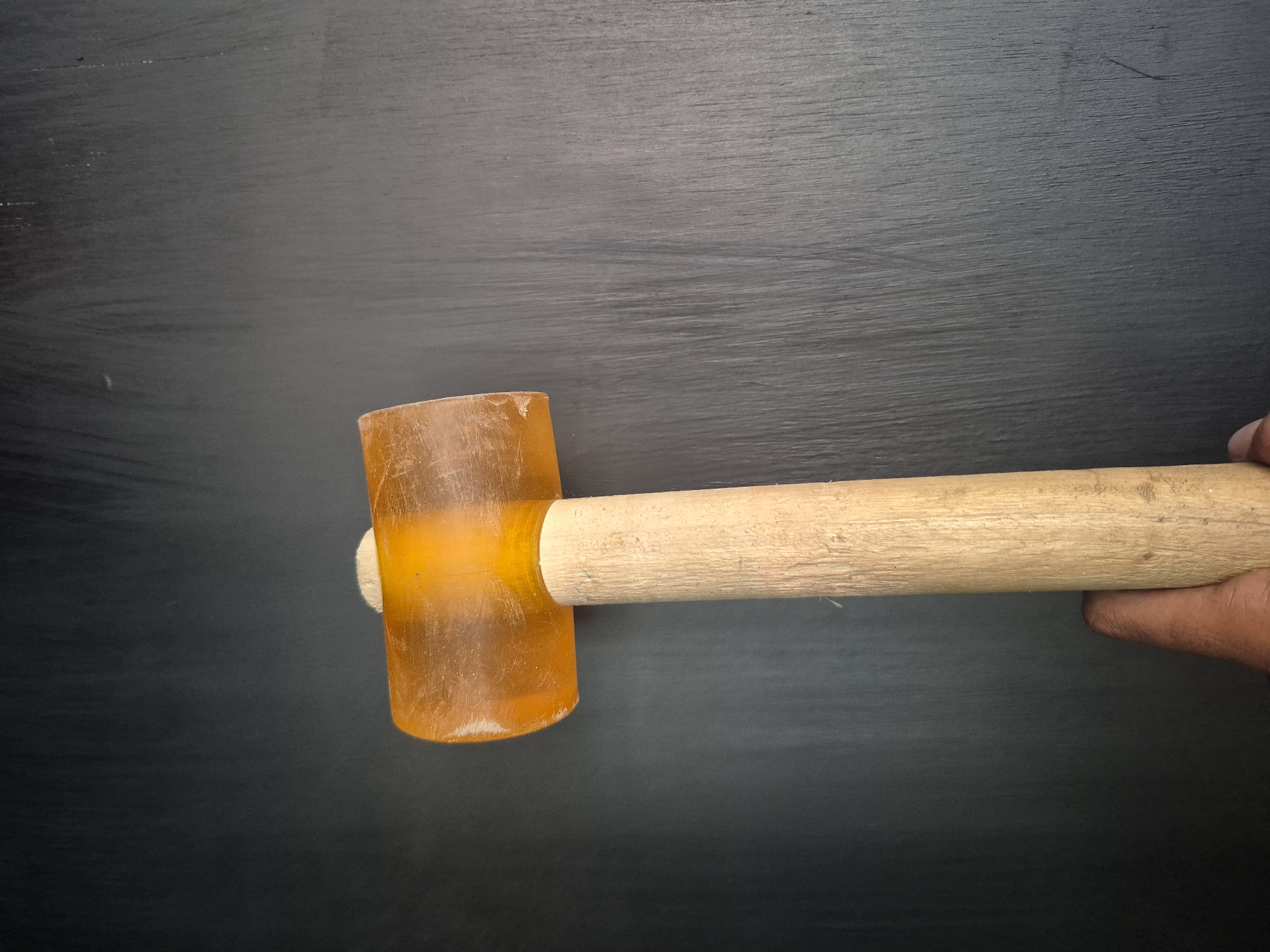 An overhead shot shows a hand holding a rubber mallet against a dark background that appears to be a chalkboard. The mallet has an orange, translucent rubber head and a light-colored wooden handle. The hand is holding the mallet near the end of the handle. The surface is dark and has subtle linear streaks, suggesting it could be a chalkboard. There is no information to specifically place this image in Lhokseumawe, Indonesia.