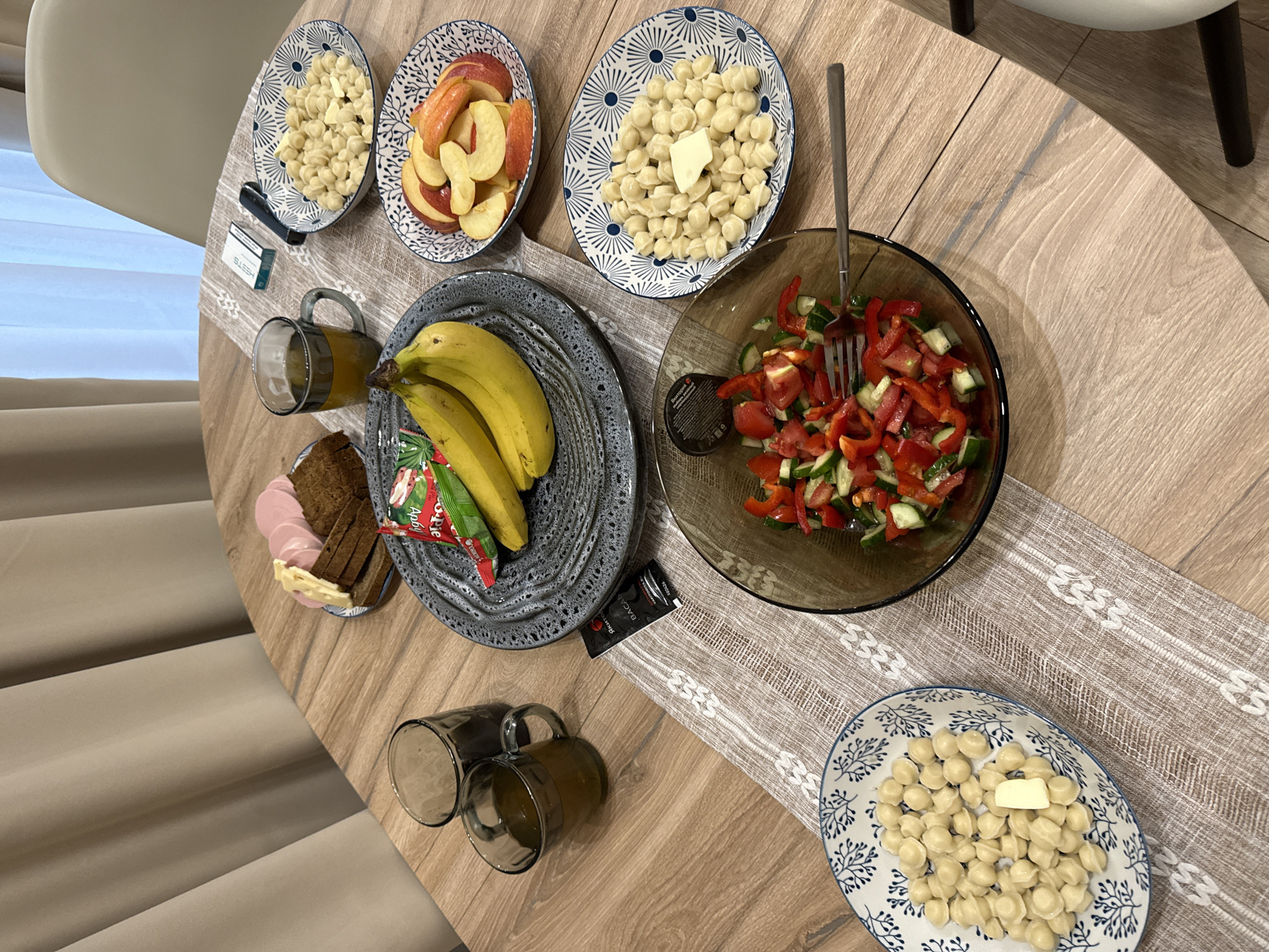 The image shows a table setting with food. The table is round and made of wood. A patterned runner lies across the table. On the table are: three plates with boiled dumplings, each with a pat of butter; a plate of sliced apples; a plate of bananas; a salad in a glass bowl; a plate with slices of bread and sausage; and two mugs with a yellowish liquid. Also visible are a pack of cigarettes and a small packet of sauce. In the background are a chair and a curtain. The location may be Buzhaninovo, Russia.