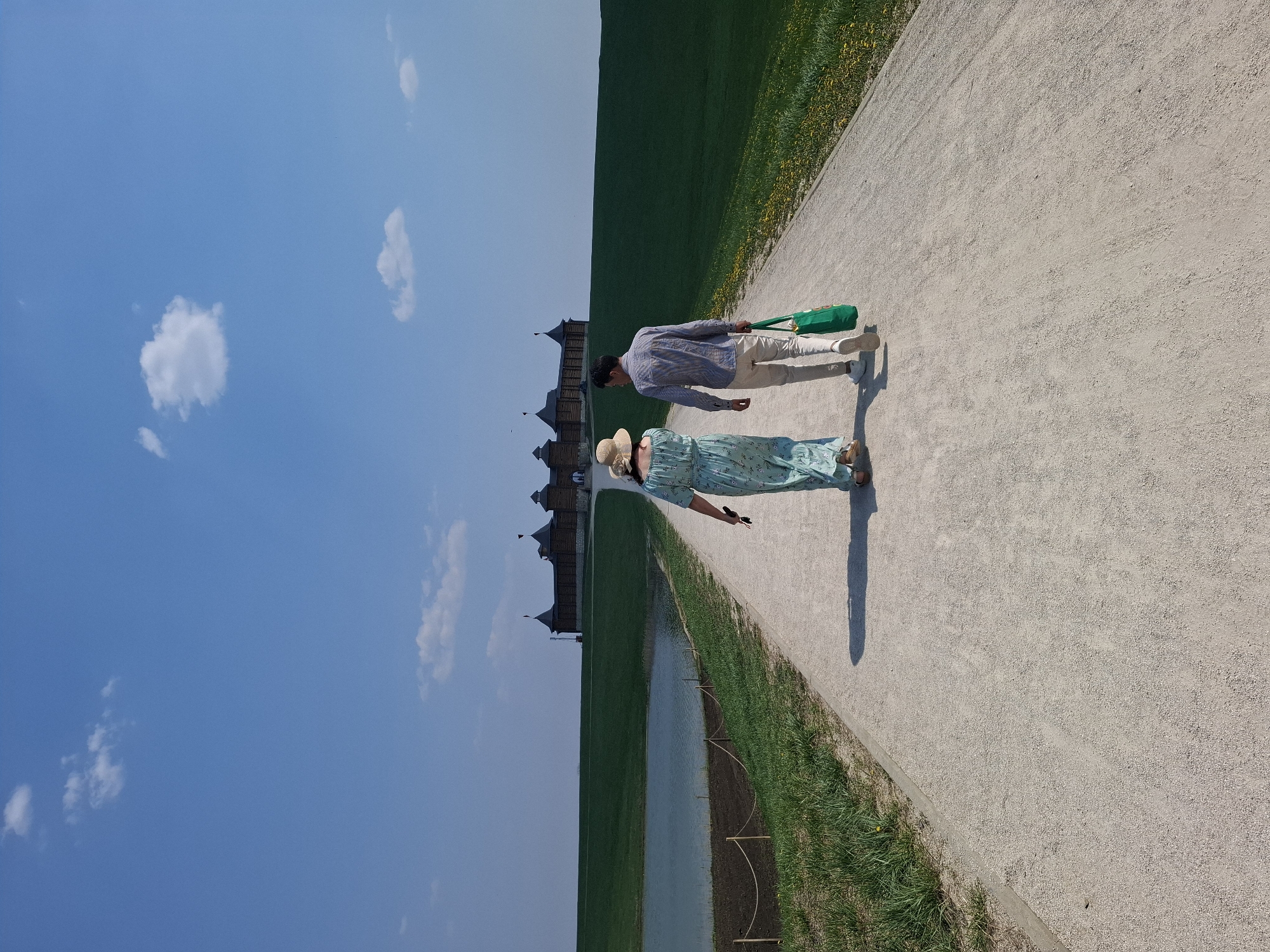 A photo taken in the city of Алисово, Russia depicts a woman and a man walking along a path next to a grassy field. The woman is wearing a light green floral dress and a straw hat. The man is wearing a light blue striped shirt, khaki pants, and sneakers. He is carrying a green tote bag. In the background, there is a wooden building with several towers. The sky is blue with a few white clouds.