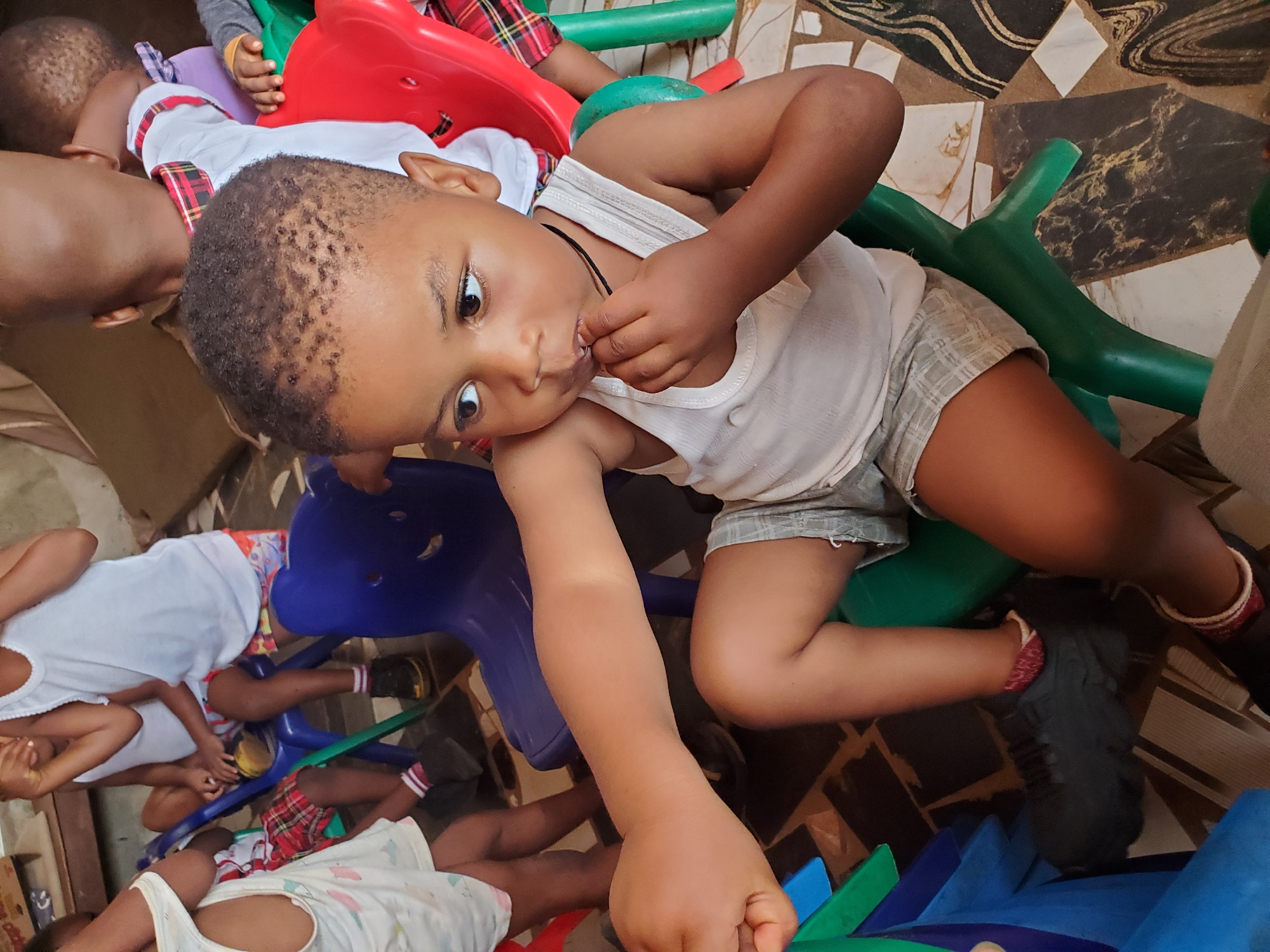 The image shows several children at what appears to be a preschool or daycare in Ukwu-Abwa, Nigeria. A young child is lying on a piece of playground equipment, possibly a plastic climbing structure or a small slide, and is holding a finger in their mouth. The child is wearing a white tank top and gray shorts. Other children are visible in the background, some wearing white shirts with plaid trim. The playground equipment is colorful, with red, blue, and green pieces. The floor has a tile pattern in various shades of brown.