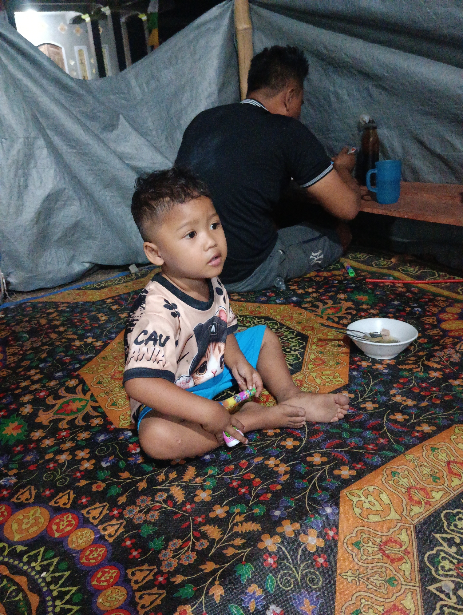 A young boy sits cross-legged on a patterned carpet, looking to his right. He wears a light-colored t-shirt with a graphic print and blue shorts. His hair is short and styled. In front of him, to his right, is a white bowl containing food and a spoon. To his left, he holds a colorful toy. Behind him, an adult man is seated facing away from the camera, wearing a black t-shirt. A blue mug and a bottle are visible on a wooden surface behind the man. The background appears to be a makeshift shelter with gray tarpaulin walls. The setting is likely a residential area in Pematang Jaya, Indonesia.