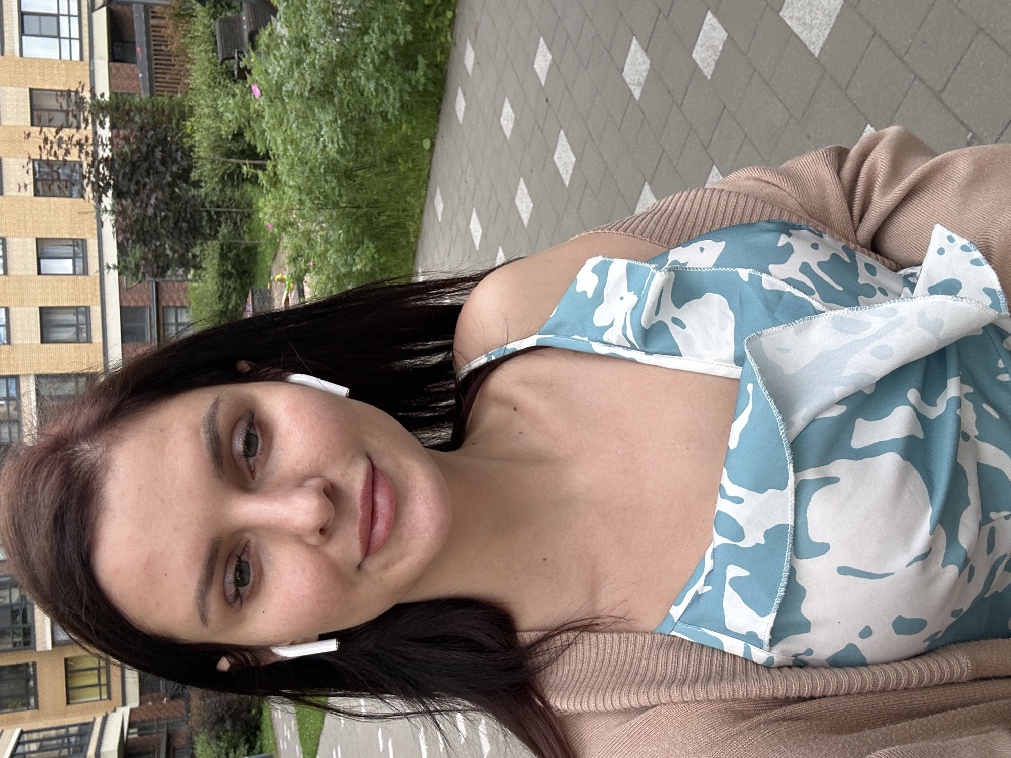 A young woman with dark hair and brown eyes is pictured in a selfie. She is wearing a light brown cardigan over a blue and white patterned top. She has white earbuds in her ears. The woman is outdoors, with a tiled walkway and lush green foliage behind her. A modern apartment building with a beige facade and dark windows is visible to the left. The lighting suggests it is daytime, possibly overcast. The woman's expression is neutral, and she is looking directly at the camera. The image is taken from a slightly upward angle. There is no visible text in the image.