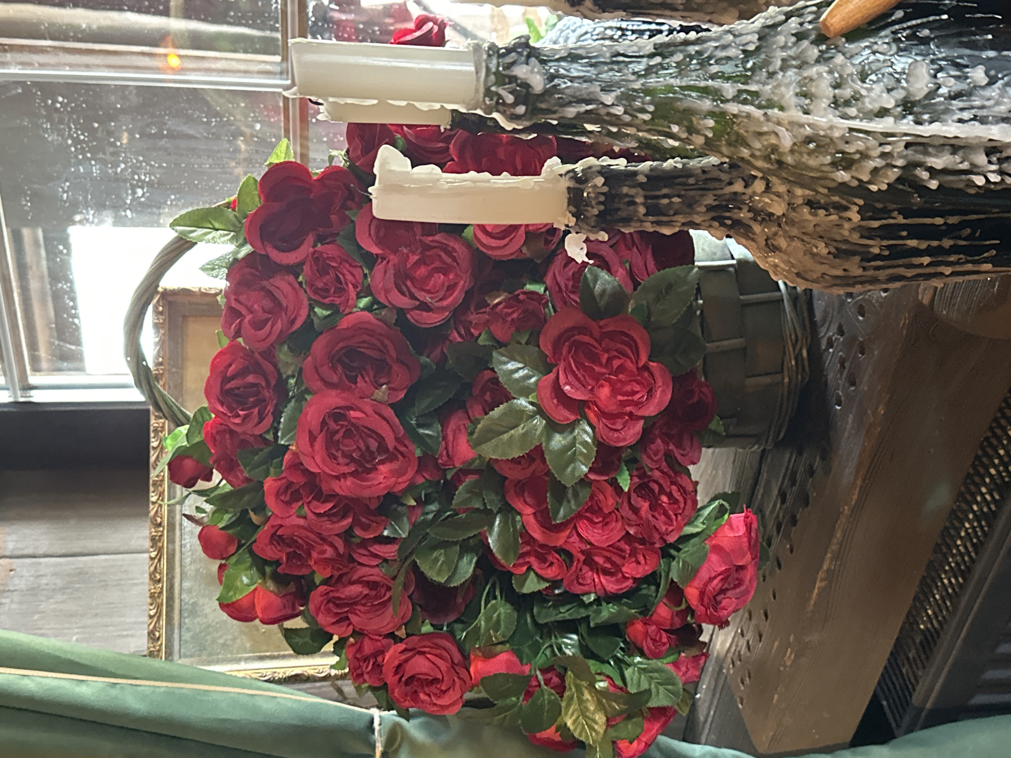 A high-angle, close-up shot shows a floral arrangement of red roses and green leaves, placed in a basket. The arrangement is set against a window with a view of the outside and a decorative mirror in a gilded frame. A wooden structure, possibly a candelabra or other decorative element, is also visible covered in what appears to be candle wax. The exact location is Новоглаголево, Russia is not clear from the image.
