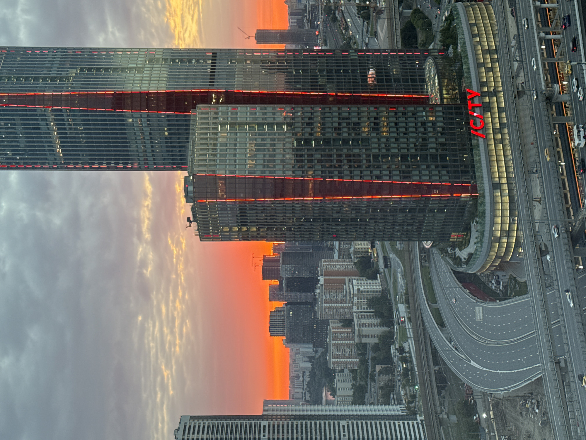 This is a medium-distance, eye-level, vertical shot of the Moscow City business district in Khimki, Russia, at sunset. The sky is a vibrant gradient of orange, pink, and gray clouds. Two prominent skyscrapers dominate the frame. The taller, thinner skyscraper on the right has a red illuminated vertical stripe. To its left is a wider, shorter skyscraper with a similar red illuminated vertical stripe and the red illuminated text "/CITY" on its circular base. The base of the shorter skyscraper has a glass facade and is surrounded by a green landscaped area. In the foreground, a multi-level highway interchange curves through the scene, with cars visible on the roadways. The surrounding urban landscape consists of numerous residential buildings and other skyscrapers extending into the distance, with a hint of a body of water on the left side of the frame. Construction cranes are visible in the background. The lighting suggests it is dusk or early evening.