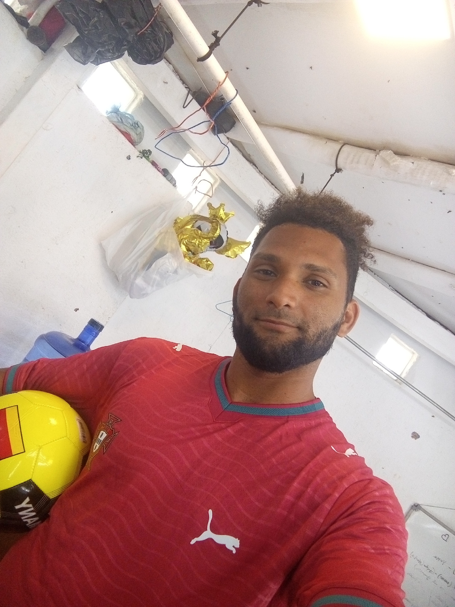 A man with curly brown hair and a beard takes a selfie in a room. He is wearing a red shirt with a blue collar and a white Puma logo. He is holding a yellow and black soccer ball. The room appears to be a storage area, with various items hanging from the ceiling and stacked on shelves. There is a large blue water bottle in the background. The room is white with some exposed pipes and wiring.
