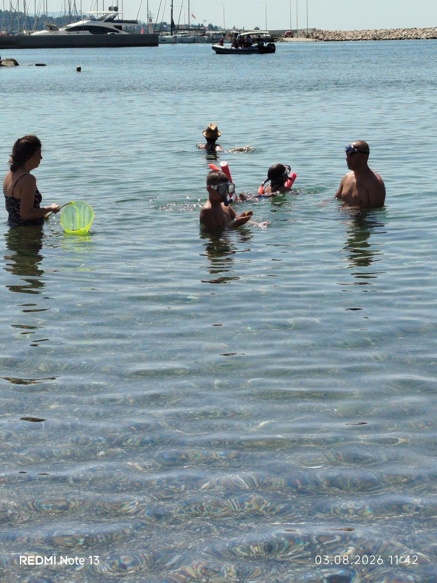 The image captures a sunny day at a coastal location, likely a harbor or bay, with people swimming in clear, shallow water. In the foreground, a woman stands in the water holding a yellow net, facing left. To her right, a young person wearing a snorkel mask and a red floatation device is visible. Further back and to the right, an adult male also wearing a snorkel mask is swimming. In the mid-ground, another person wearing a straw hat is visible, partially submerged. The background reveals a harbor with several boats, including a large yacht and smaller vessels, docked along a stone jetty. A rocky shoreline is also visible in the distance, with some structures and trees. The water is calm with gentle ripples, reflecting the sunlight and revealing the sandy seabed. The timestamp "03.08.2026 11:42" and the text "REDMI Note 13" are present in the lower corners of the image. The overall scene depicts a leisurely activity in a picturesque seaside setting, suggesting a tourist or recreational area within the Sithonia Municipal Unit of Greece.