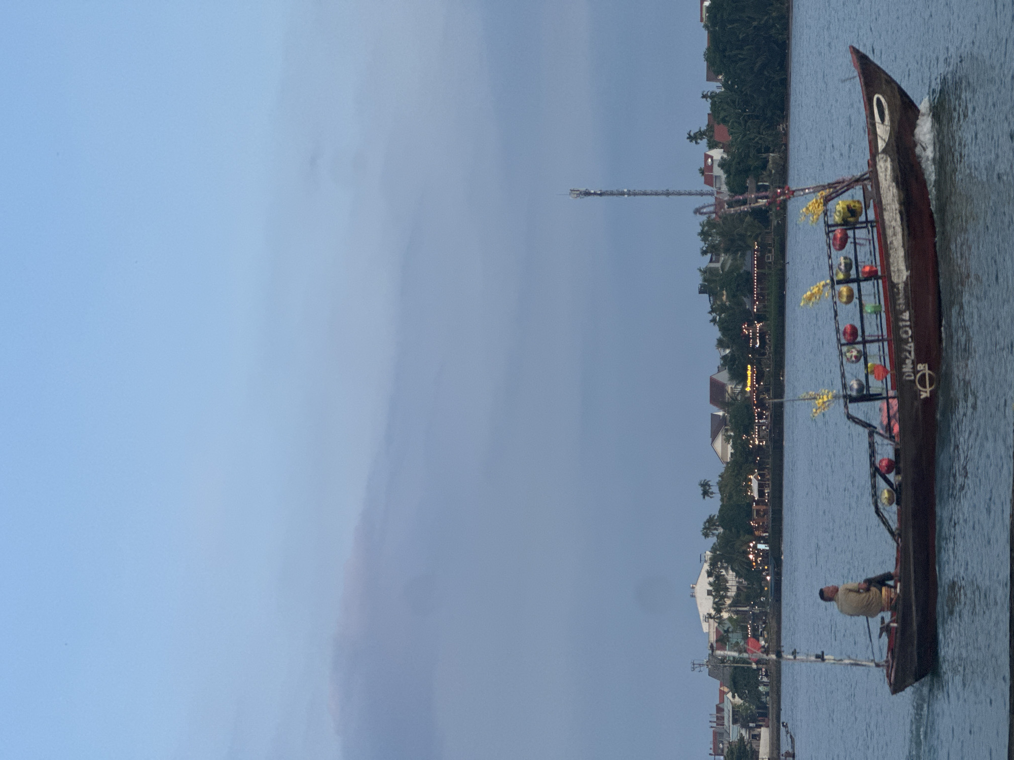 A medium shot depicts a boat, operated by a man, traveling on a body of water adjacent to the city of Hội An Ward, Vietnam. The boat, adorned with multi-colored spherical objects and yellow decorations, is moving from the bottom of the frame towards the top. The man, situated at the back of the boat, appears to be steering or rowing. The shoreline is populated with buildings, trees, and a tall communications tower. The sky is overcast, indicating either early morning or late afternoon. The environment suggests a water channel passing through a populated area.