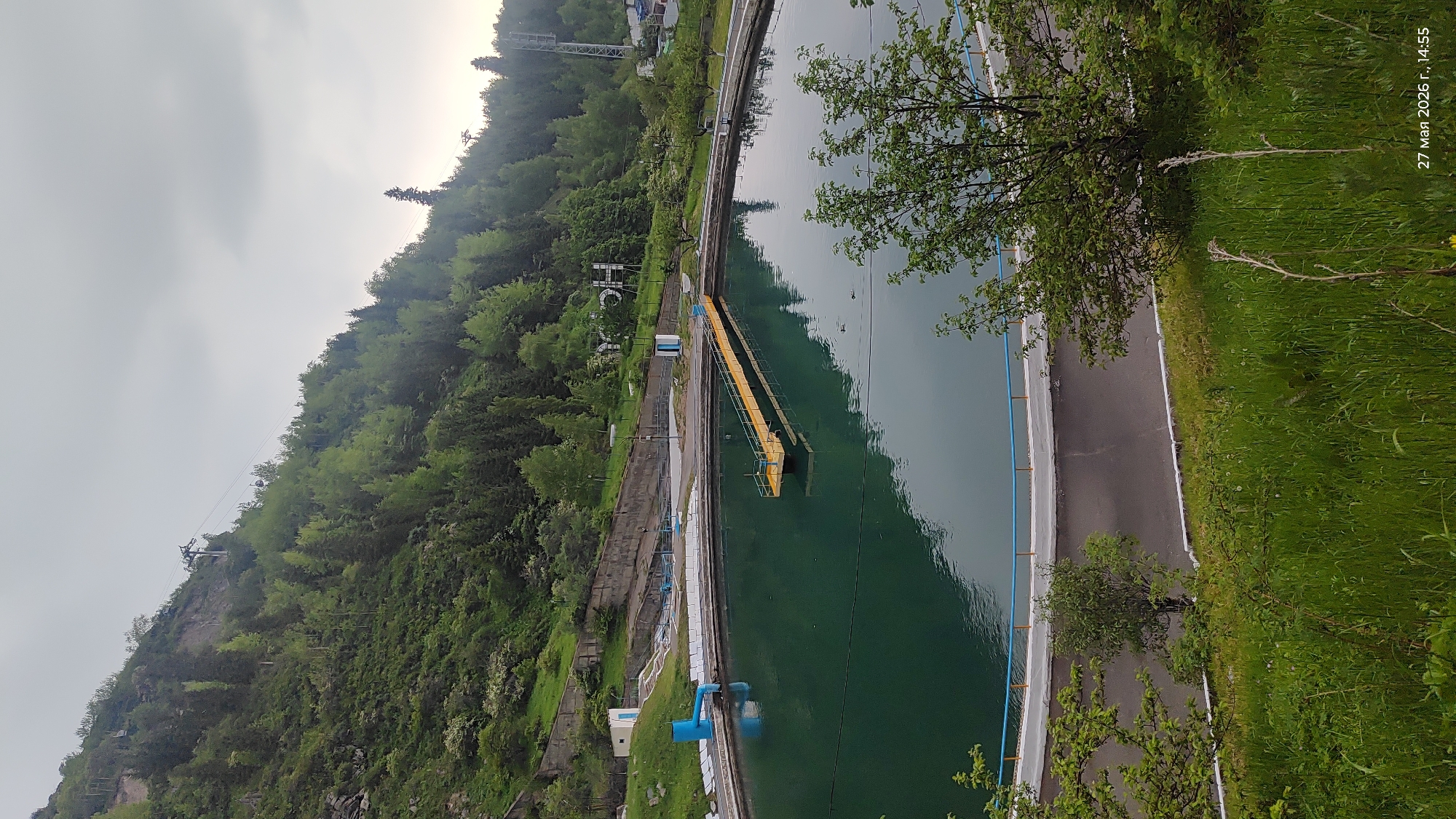 The image depicts a water reservoir located in the mountains near Almaty, Kazakhstan. The reservoir is long and narrow, with a green-tinted water surface. A yellow bridge-like structure extends into the water from the bank. A road runs along one side of the reservoir, bordered by green grass and some trees. On the opposite side, a steep, tree-covered mountain rises up. A cable car or ski lift is visible ascending the mountain. The sky is overcast, creating soft lighting. A date stamp "27 мая 2026 г., 14:55" (May 27, 2026, 14:55) is present in the upper right corner of the image.
