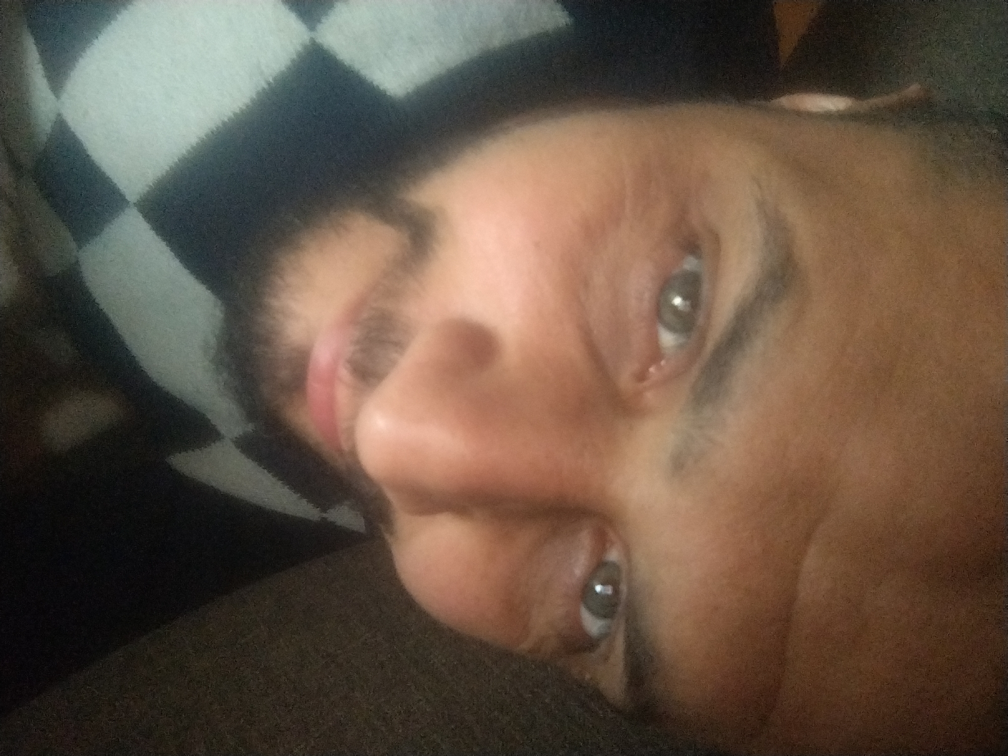 The image shows a close-up, overhead view of a person's face. The person appears to be lying down, with their head tilted to the side. Their left eye, which is looking directly at the camera, is visible and appears to be gray or green. The person has a light complexion and appears to have short, dark hair. They are wearing a black and white checkered garment. The background is dark and out of focus. The scene is indoors, possibly in a home environment.