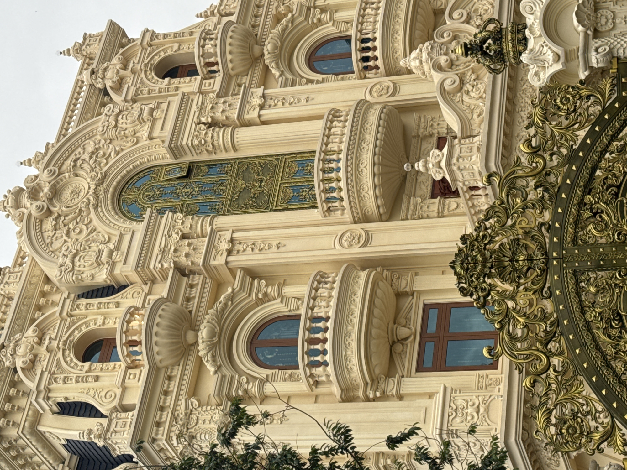 The image depicts the ornate exterior of a lavish building, likely a private residence or a luxury hotel, in Da Lat, Vietnam. The architecture is characterized by extensive decorative plasterwork in a pale yellow hue, featuring intricate floral motifs, scrolls, and classical architectural elements such as columns and balustrades. Several balconies with rounded profiles and elaborate railings punctuate the facade. The windows are predominantly arched and framed with ornate molding, with one large arched window in the center displaying a dark, intricate metal grille over blue glass. The building's roofline is visible in the upper left, showing dark gray tiling and decorative finials. In the foreground, a section of a large, ornate golden gate is visible, adorned with elaborate scrollwork and floral patterns, suggesting a grand entrance. A few branches of dark green foliage from a tree are also present on the left side of the image, adding a touch of natural contrast to the man-made opulence. The sky above is overcast and a pale gray. There are no people visible in the image.