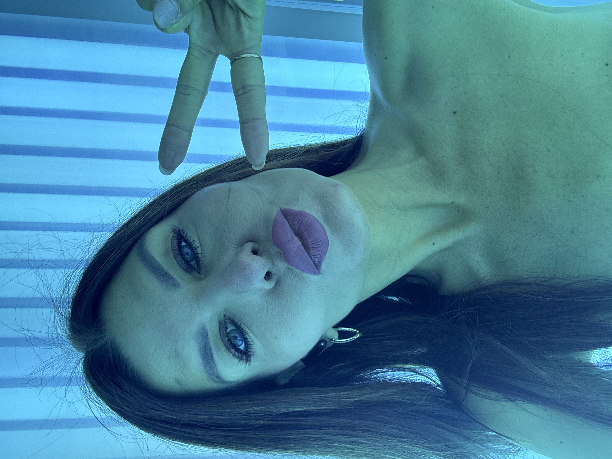 A woman lies in a tanning bed, making a peace sign with her right hand. She has long brown hair, blue eyes, and purple lipstick. She is wearing a ring on her right ring finger. The tanning bed has horizontal blue lights. The location is not discernible from the image.