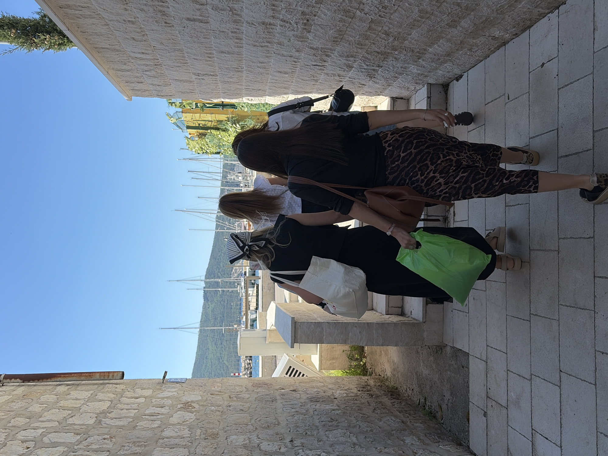 The image shows a group of three women walking along a cobblestone path in Meljine, Montenegro. The path is bordered by stone walls on both sides. The first woman is wearing animal-print pants, a black top, and a brown shoulder bag. The second woman is wearing a white top. The third woman is wearing a black dress and carrying a white tote bag and a green shopping bag. In the background, a harbor with sailboats and a tree-covered hill can be seen. The sky is blue.