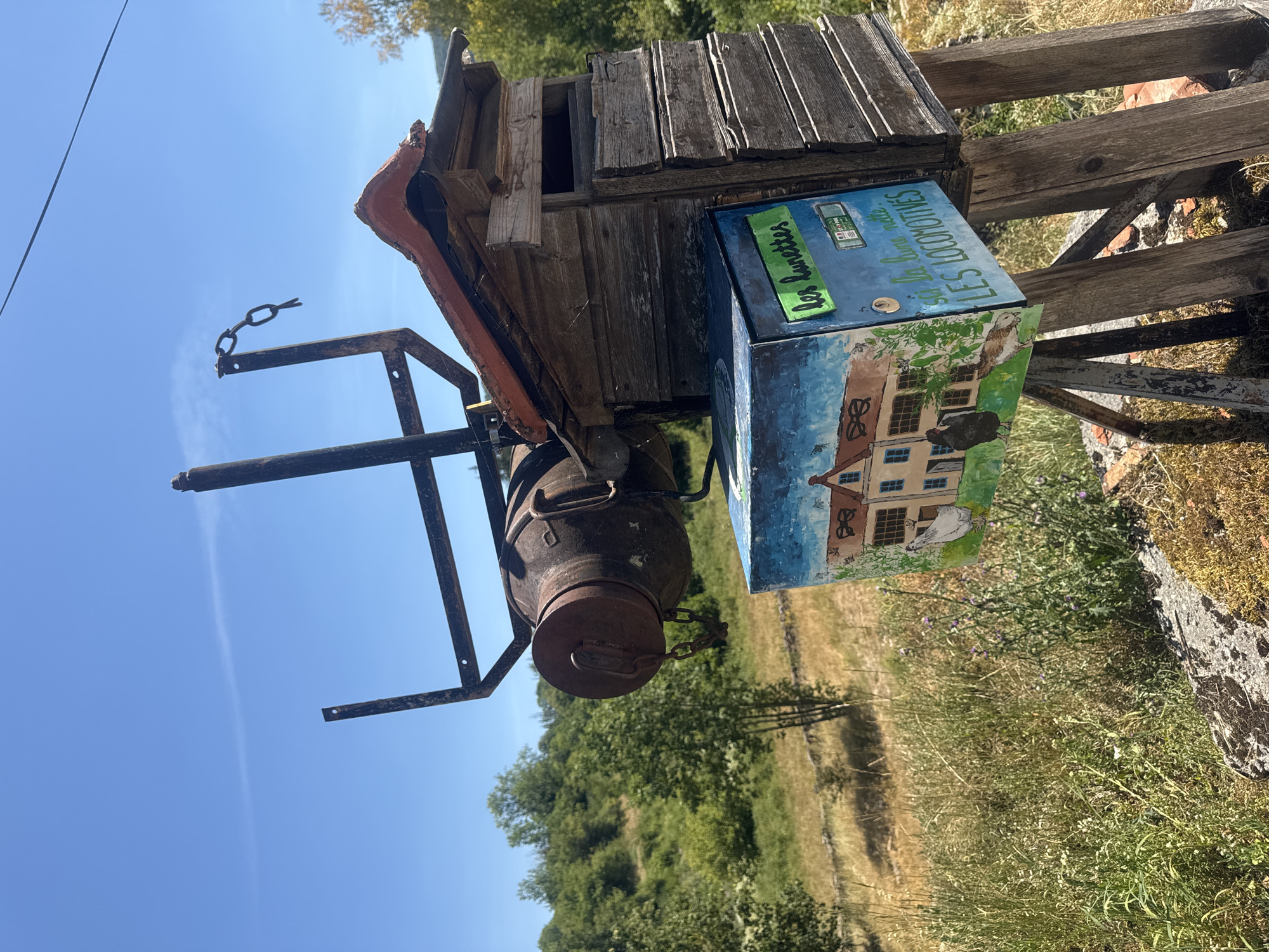 This is an outdoor, eye-level, full shot of a rustic, handmade mailbox situated on a hillside in Coteaux du Lizon, France. The mailbox is a multi-component structure. The main body appears to be constructed from weathered, horizontal wooden planks, topped with a section of red tile roofing. Attached to the top is a rusty metal frame with a chain hanging from it, and a large, rusty, cylindrical object resembling a boiler or industrial part suspended by the frame. A smaller, square box, painted with a pastoral scene of a house and chickens, is attached to the front of the wooden structure. The box has text written on it that reads "les lunettes" and "si la lune naît LES LOCOMOTIVES". The background consists of a dry, grassy hillside with scattered trees under a clear blue sky with a faint wispy cloud. The scene is well-lit by natural sunlight. There are no people or animals visible in the frame other than those depicted in the painting on the mailbox.