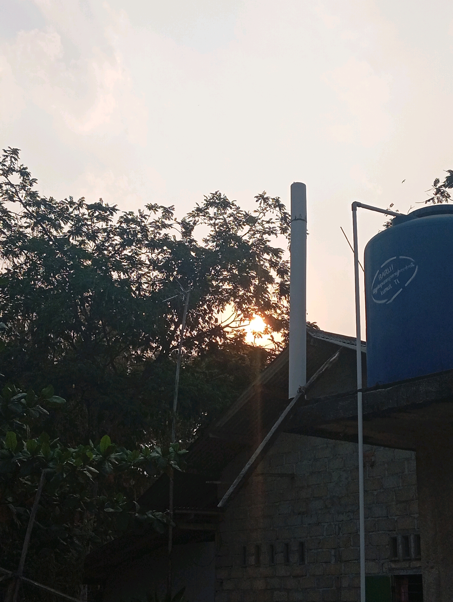 The image captures a sunset scene in Leman Benawi, Indonesia. The sky is predominantly pale orange and white, with some wispy clouds. In the foreground, a large tree with dense green foliage dominates the left side of the frame, partially obscuring the sun, which is a bright, diffused orb. Behind the tree, a house with a dark, angled roof is visible. A white cylindrical pipe, possibly a chimney or ventilation shaft, stands vertically to the right of the house. Further to the right, a large, blue water tank is perched atop a brick structure. The brickwork of the structure is visible, with several small, dark rectangular openings. A white pipe runs vertically along the side of the water tank. The overall impression is one of a rural or semi-rural setting during the late afternoon or early evening. No people are visible in the image.