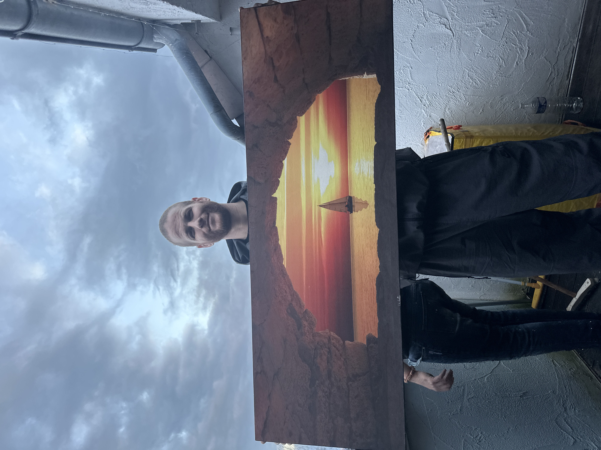 The image appears to be taken from a low angle, looking upwards, possibly from a balcony or rooftop. A person with a short beard is holding a painting of a seascape featuring a sunset and a sailboat framed by what looks like a rocky cave opening. The sky behind the person is overcast. In the background, there's a white wall with rough texture, a yellow object, and a transparent water bottle. The photograph was taken in an environment with an apparent view of the sky and possible architectural structures above. There is no clear way to verify that the photo was taken in Heinebach, Germany.