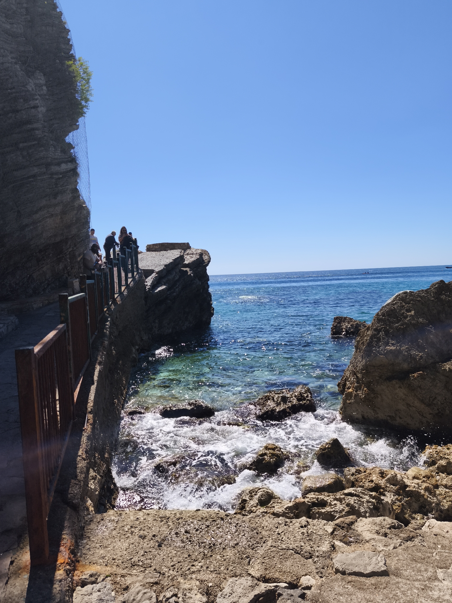 A coastal scene shows a path along a rocky cliff leading to a group of people overlooking the sea. The path is bordered by a weathered wooden railing. The clear turquoise sea laps against the rocky shore. In the distance, small boats are visible on the horizon. The sky is clear and blue. The location is Unknown, Unknown.