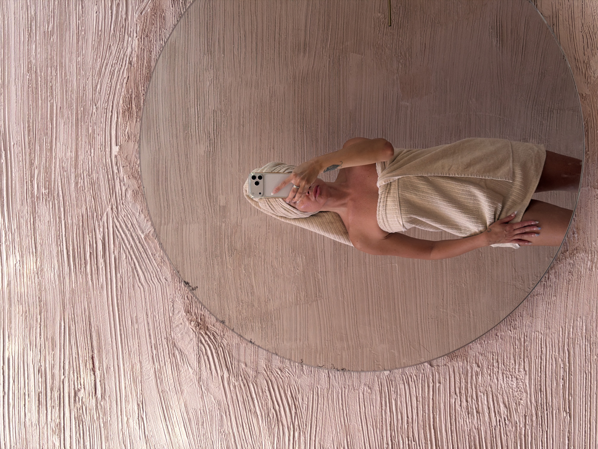 A woman wrapped in a towel and wearing a towel on her head is taking a selfie in a round mirror. The mirror is mounted on a wall with a textured, pinkish-beige finish. The wall texture appears to be thick, with vertical lines. The woman is holding a white smartphone with multiple camera lenses. The mirror's reflection shows a partial view of her body, including her shoulders, arms, and legs. This picture may have been taken in a bathroom located in North Kuta, Indonesia.