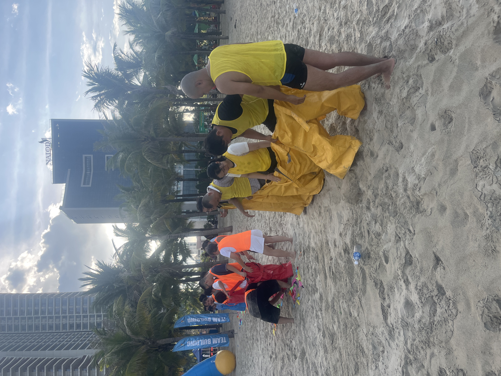 A group of people are on a sandy beach in Da Nang, Vietnam, participating in a team-building activity. Most of the participants are wearing yellow or orange vests. Some are struggling to get inside large yellow or red sacks, while others are standing by, observing or helping. In the background, there are palm trees and tall buildings, characteristic of the coastal city's skyline. The sky is cloudy with sunlight breaking through. The beach is littered with small, colorful pieces of paper and a plastic water bottle. The overall scene depicts an outdoor recreational event in a tropical urban setting.
