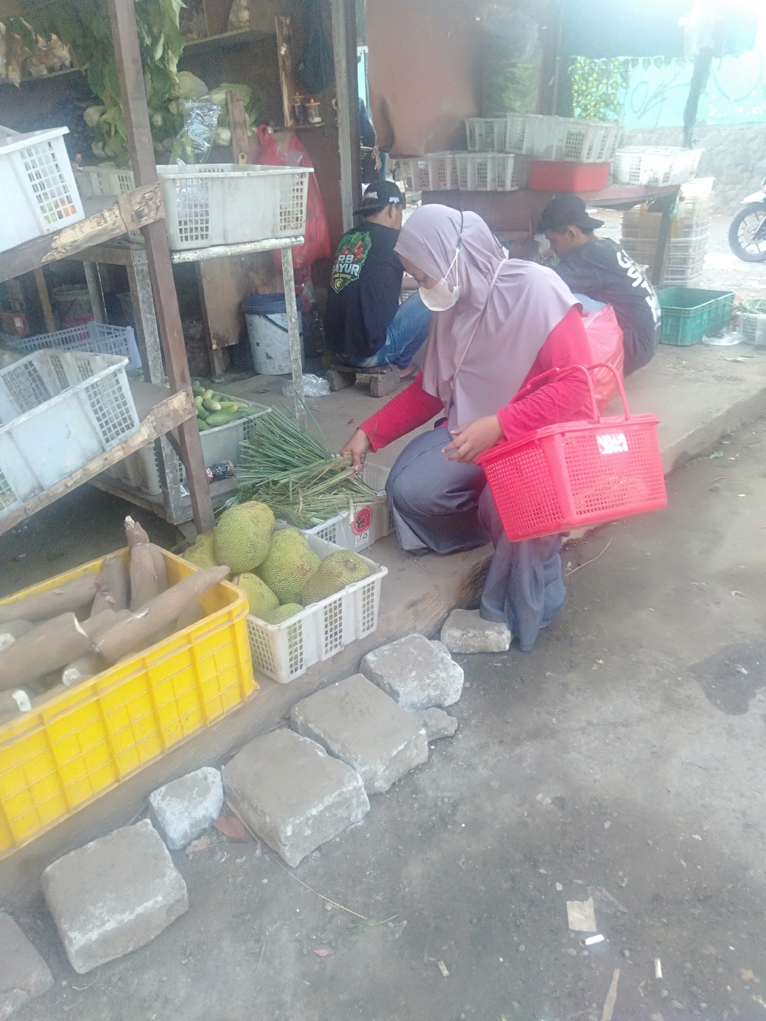 The image depicts an outdoor market scene in Sukadami, Indonesia. In the foreground, a woman wearing a purple hijab and a face mask is holding a red basket. She is seated on the ground and appears to be interacting with a selection of produce. To her left, a yellow crate contains several large, brown root vegetables, possibly cassava. Next to this, a white basket is filled with green jackfruit. Behind the woman, a stand displays more produce, including what appears to be cucumbers in a white basket. Several white plastic crates are stacked on shelves in the background, some containing vegetables. To the right of the woman, a young person wearing a black t-shirt with white lettering and a black baseball cap is also seated, facing away from the camera. The ground is a mixture of dirt and concrete, with several large concrete blocks scattered around. The overall environment suggests a bustling marketplace with fresh produce being sold.